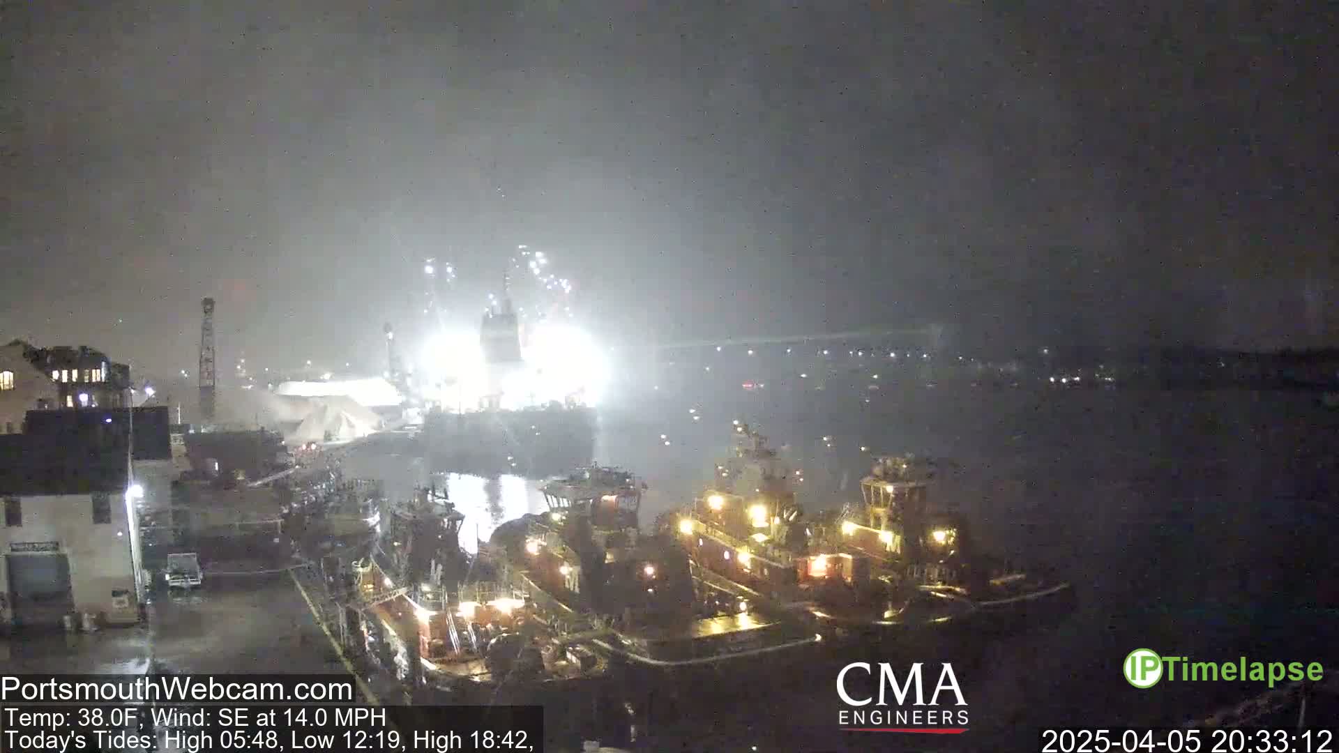 At night, under hazy conditions, several tugboats are moored near a brightly lit dock where a large vessel is undergoing operations.