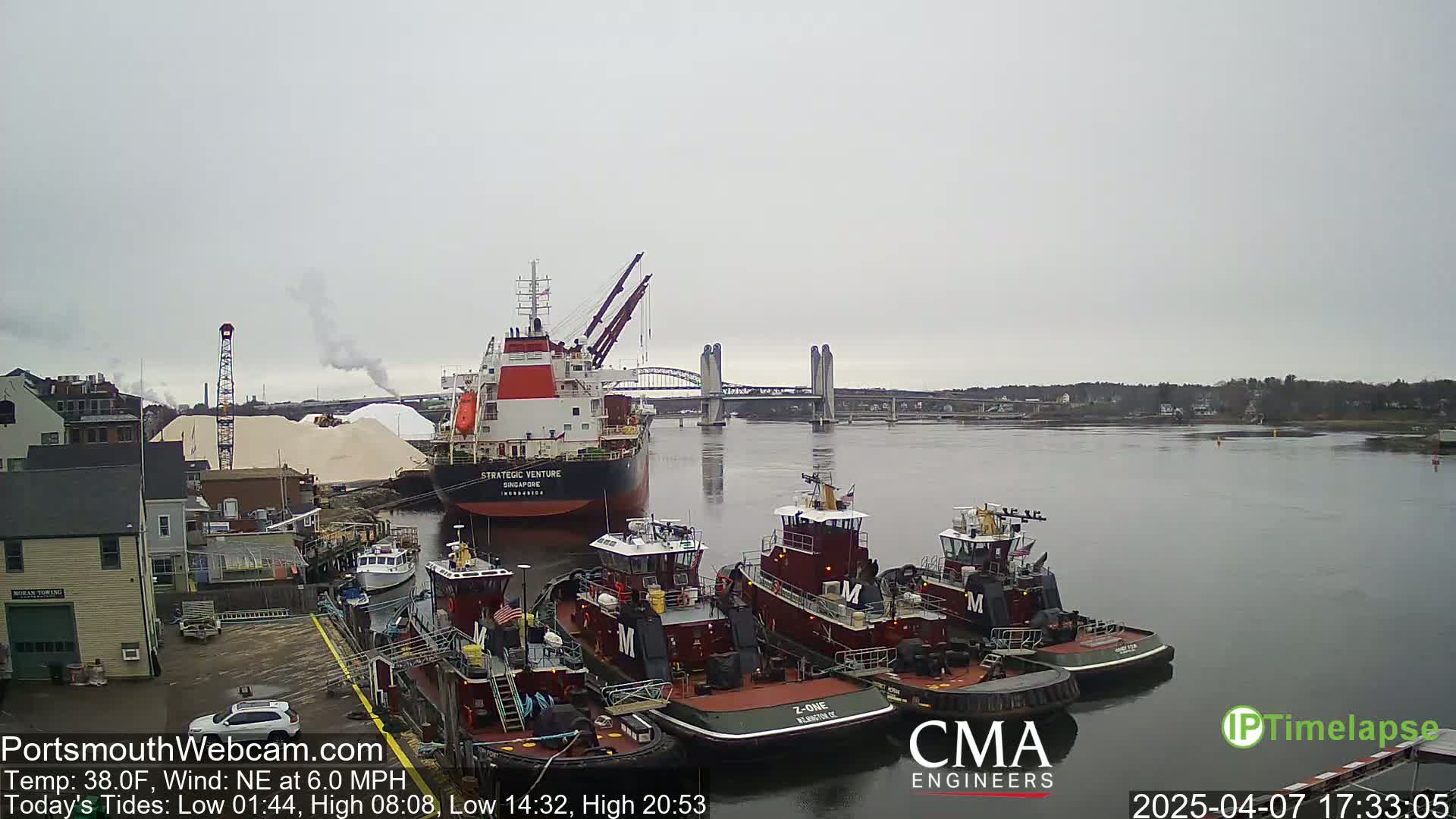 A large cargo ship is docked near several tugboats, all in a harbor under an overcast sky.