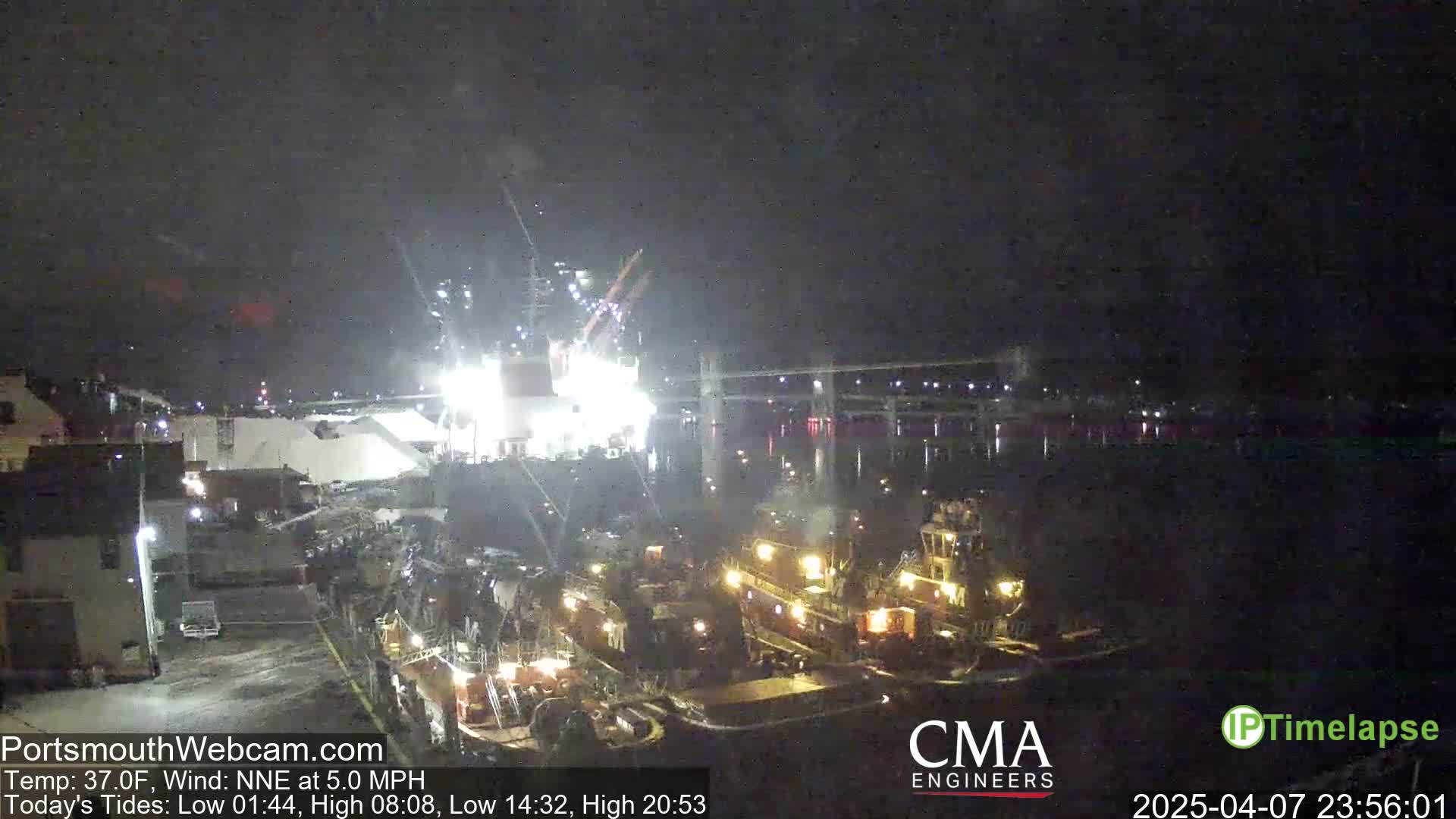 At night, under a dark sky, several tugboats are moored near a brightly lit industrial area with a large pile of white material, and a bridge is visible in the distance.