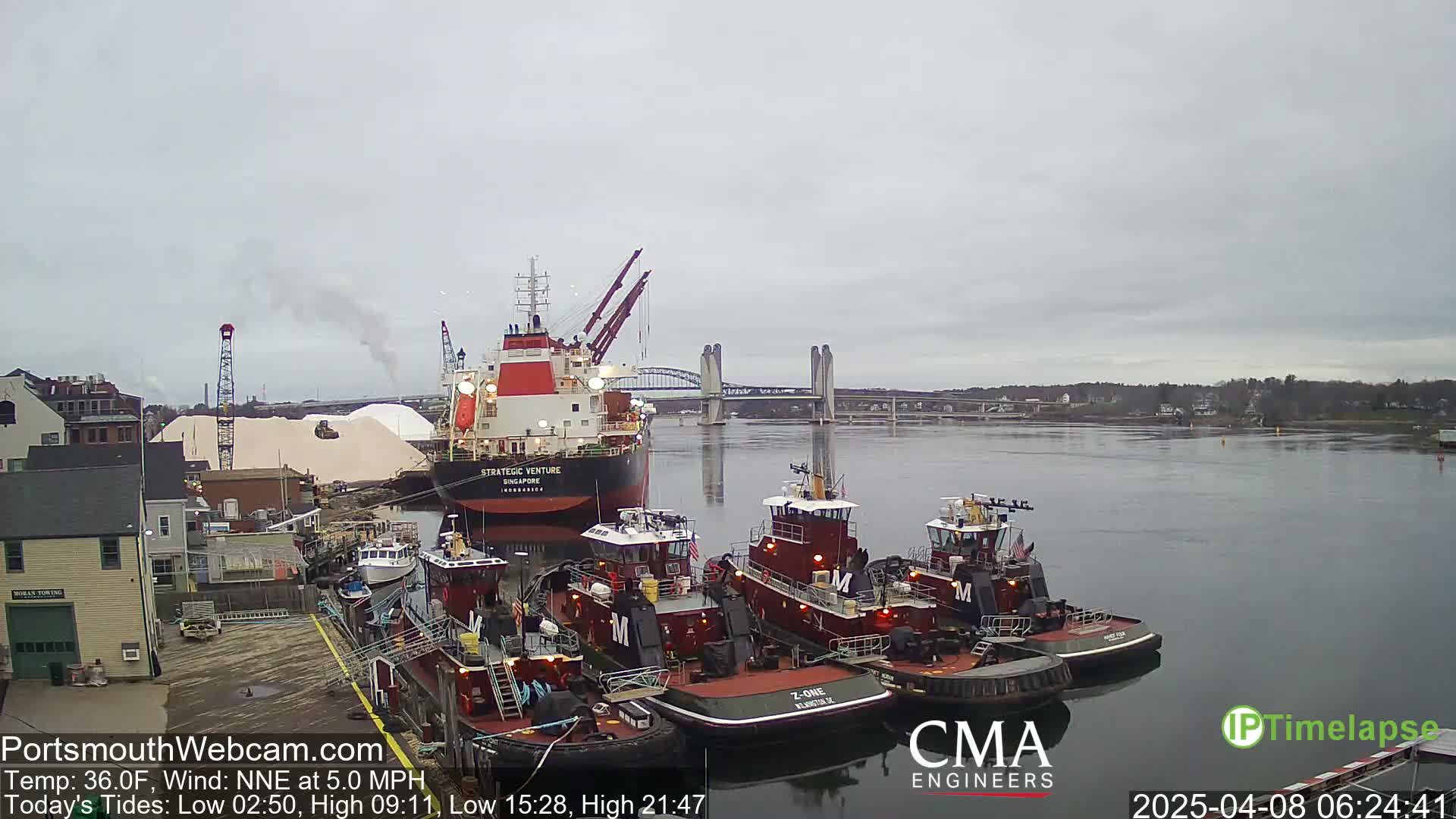 A large cargo ship is docked alongside several tugboats in a harbor under an overcast sky.