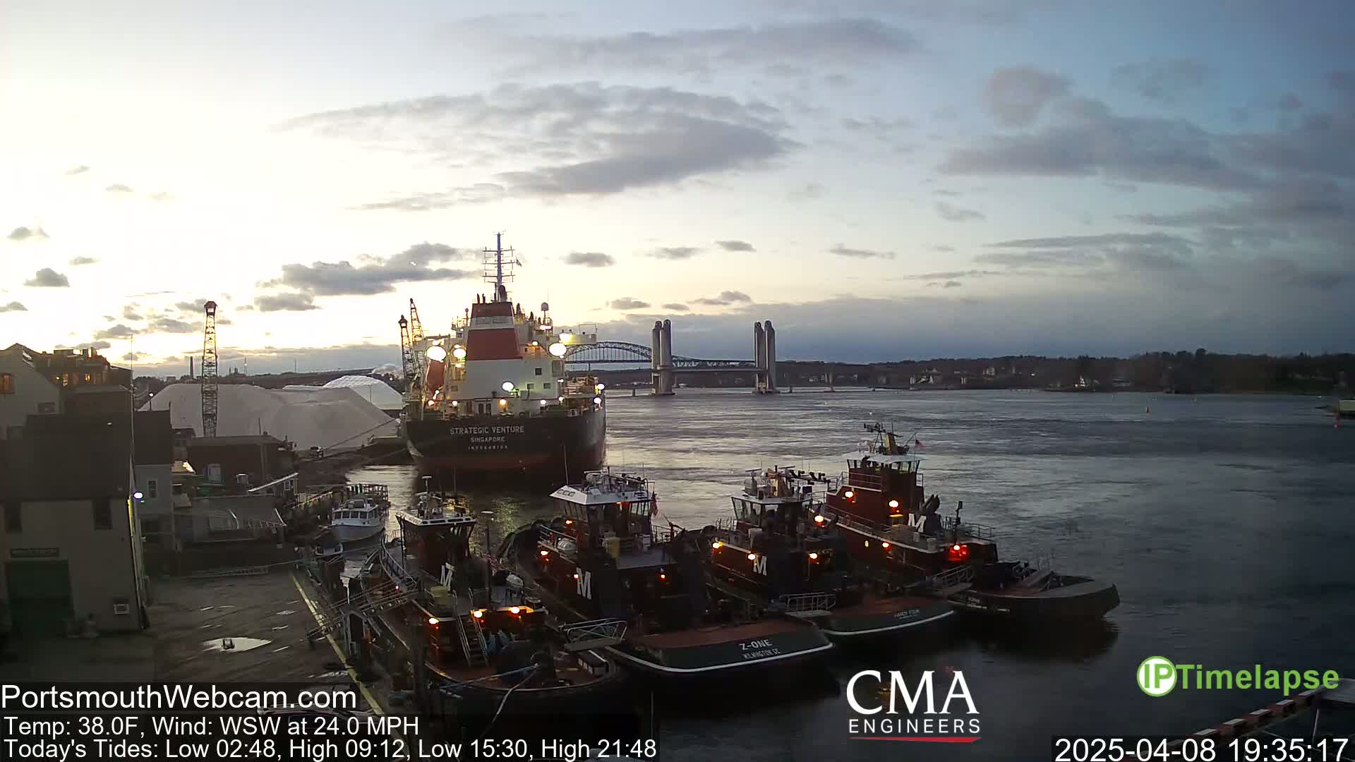 A large cargo ship is moored next to several tugboats in a harbor at dusk under a cloudy sky, with a bridge visible in the distance.