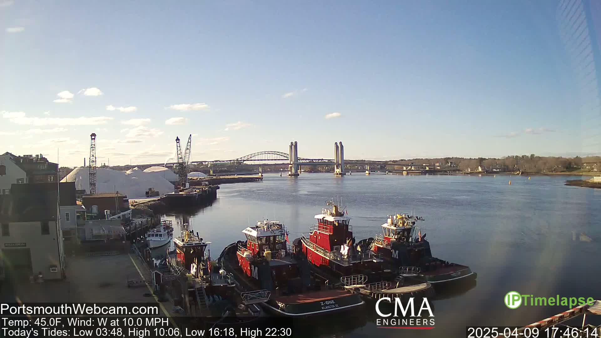 Several tugboats are moored at a dock next to a large pile of salt, with a lift bridge spanning a calm river in the background under a mostly clear sky.