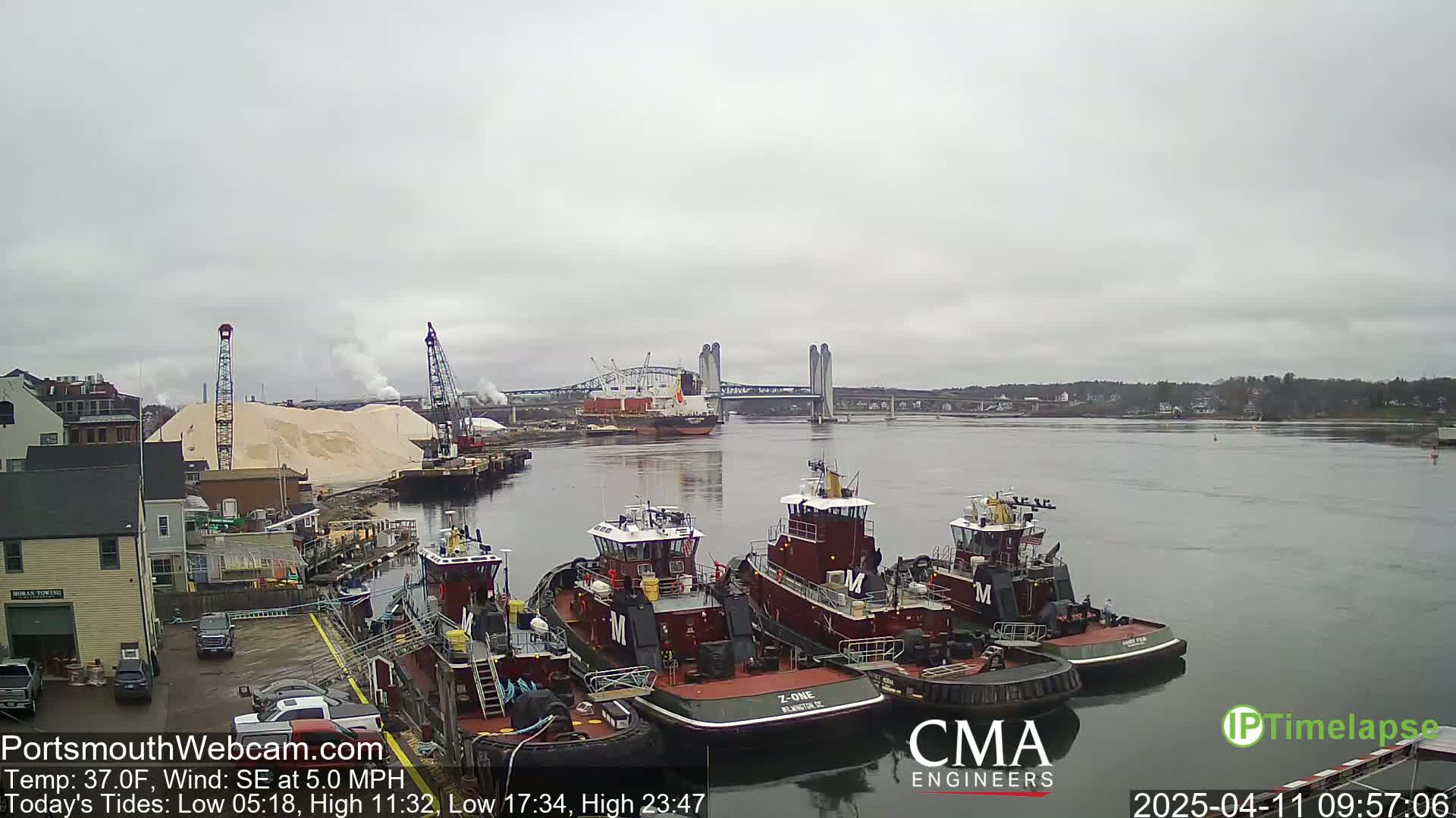 Several tugboats are moored at a dock beside a large pile of sand, with a bridge and a cargo ship visible in the distance on an overcast day.