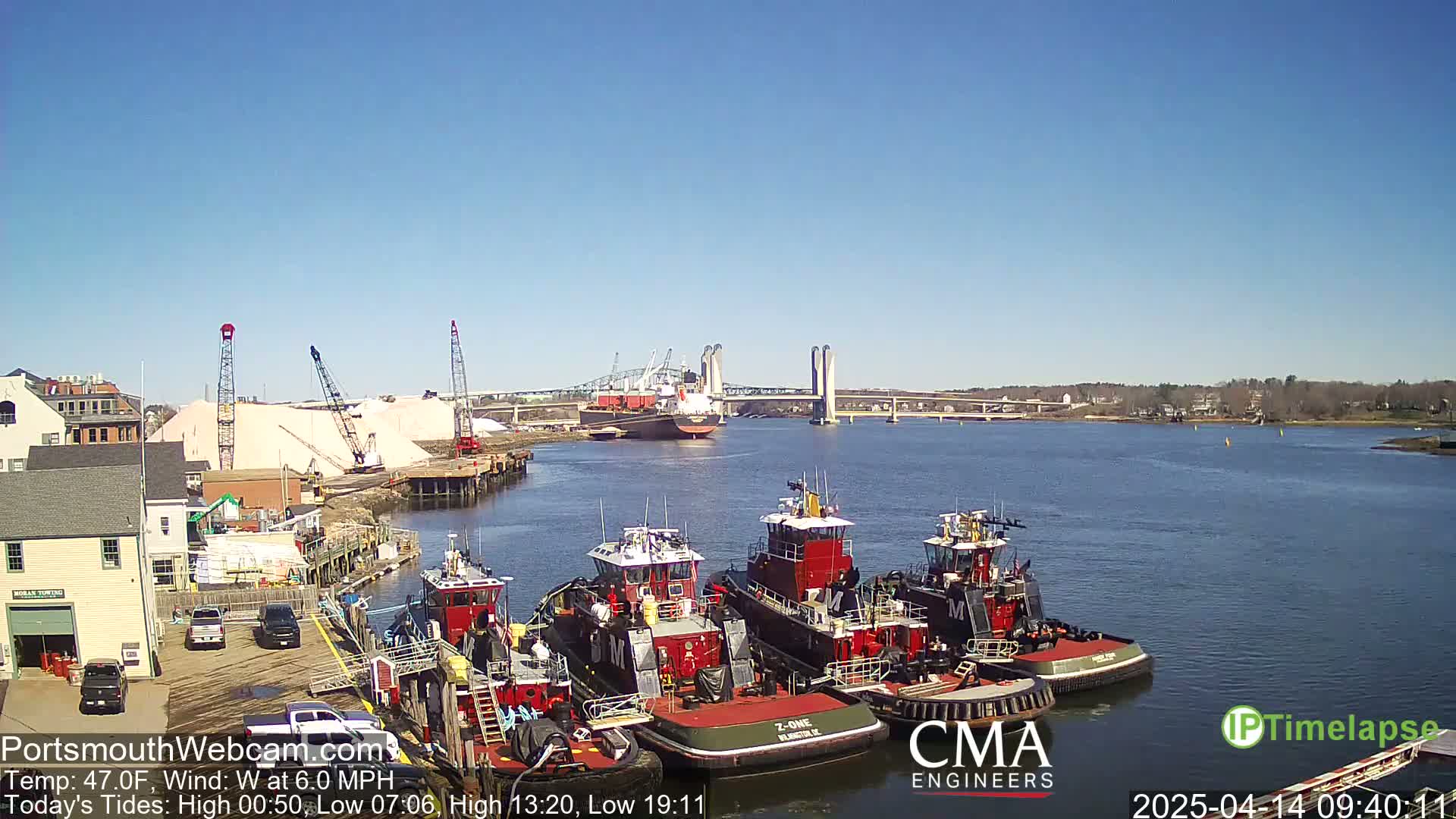Several tugboats are moored at a dock near a large pile of salt, with a cargo ship and a bridge visible in the distance on a sunny day with a light westerly wind.