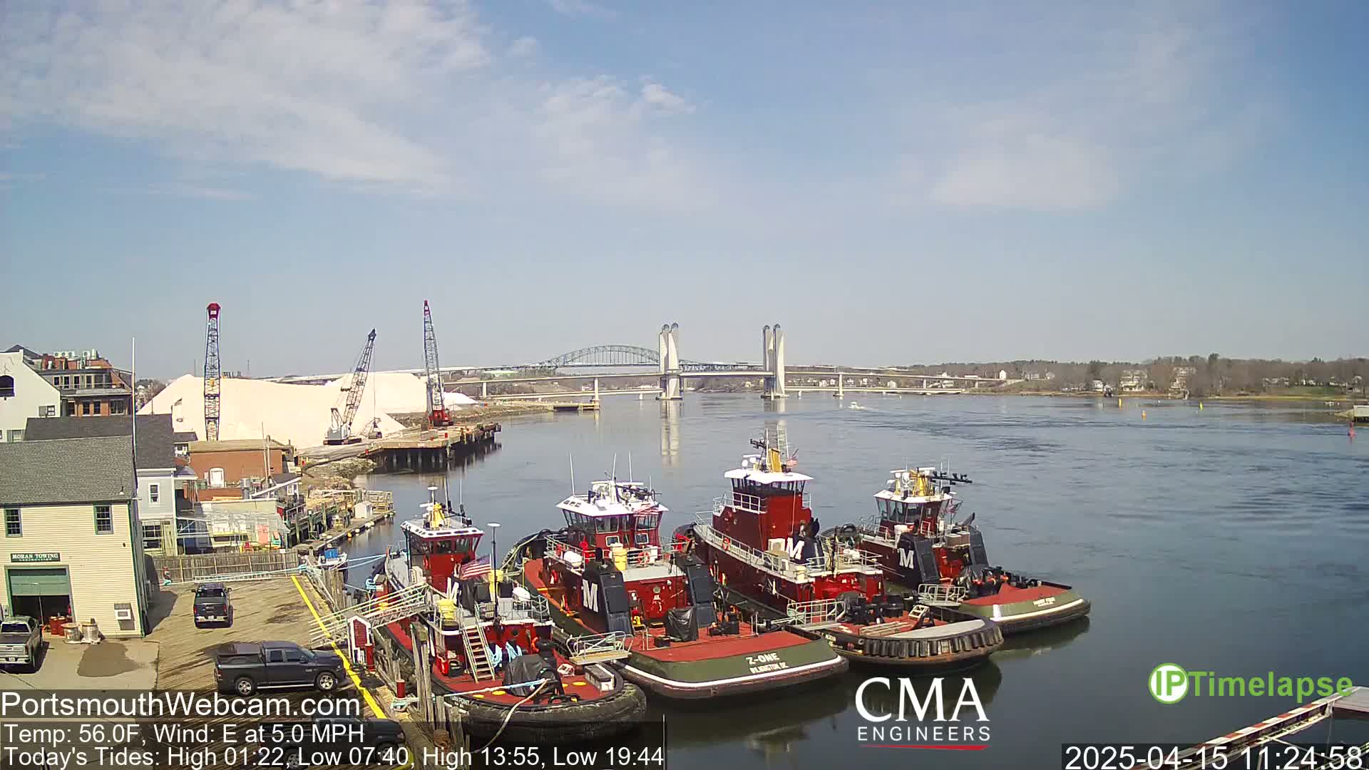 Several tugboats are moored at a dock near a large pile of salt, with a bridge in the background under a partly cloudy sky.