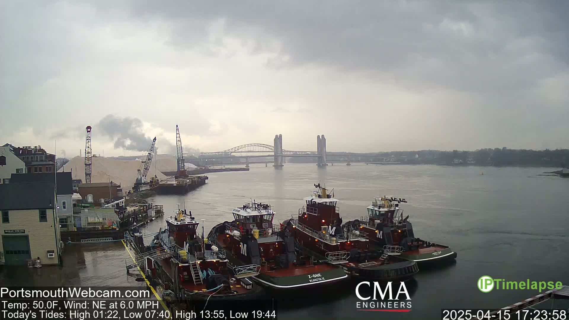 Several tugboats are moored at a dock, with a bridge and a large pile of sand visible across a calm, overcast river.