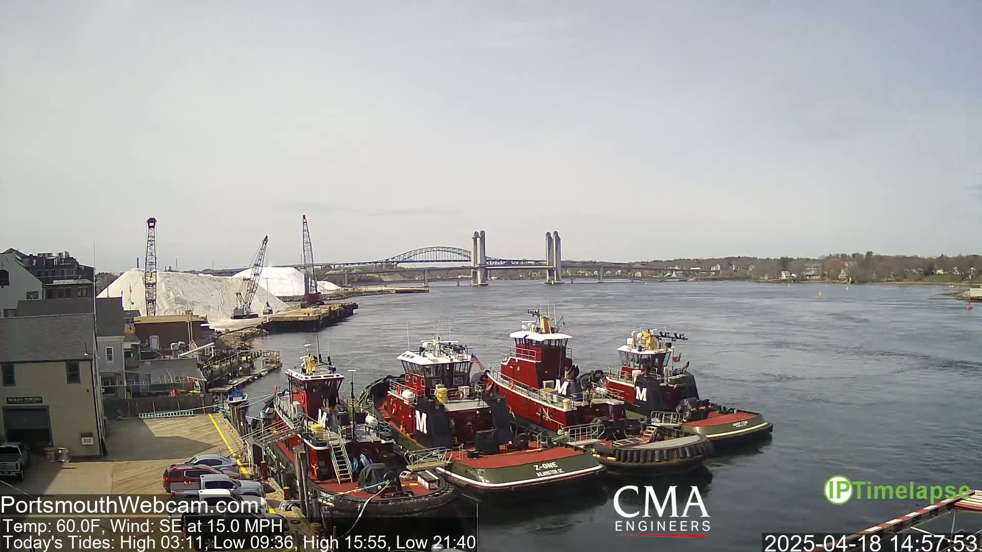 Several tugboats are docked near a large pile of white material, with a bridge in the background on a partly sunny day with a southeast wind.