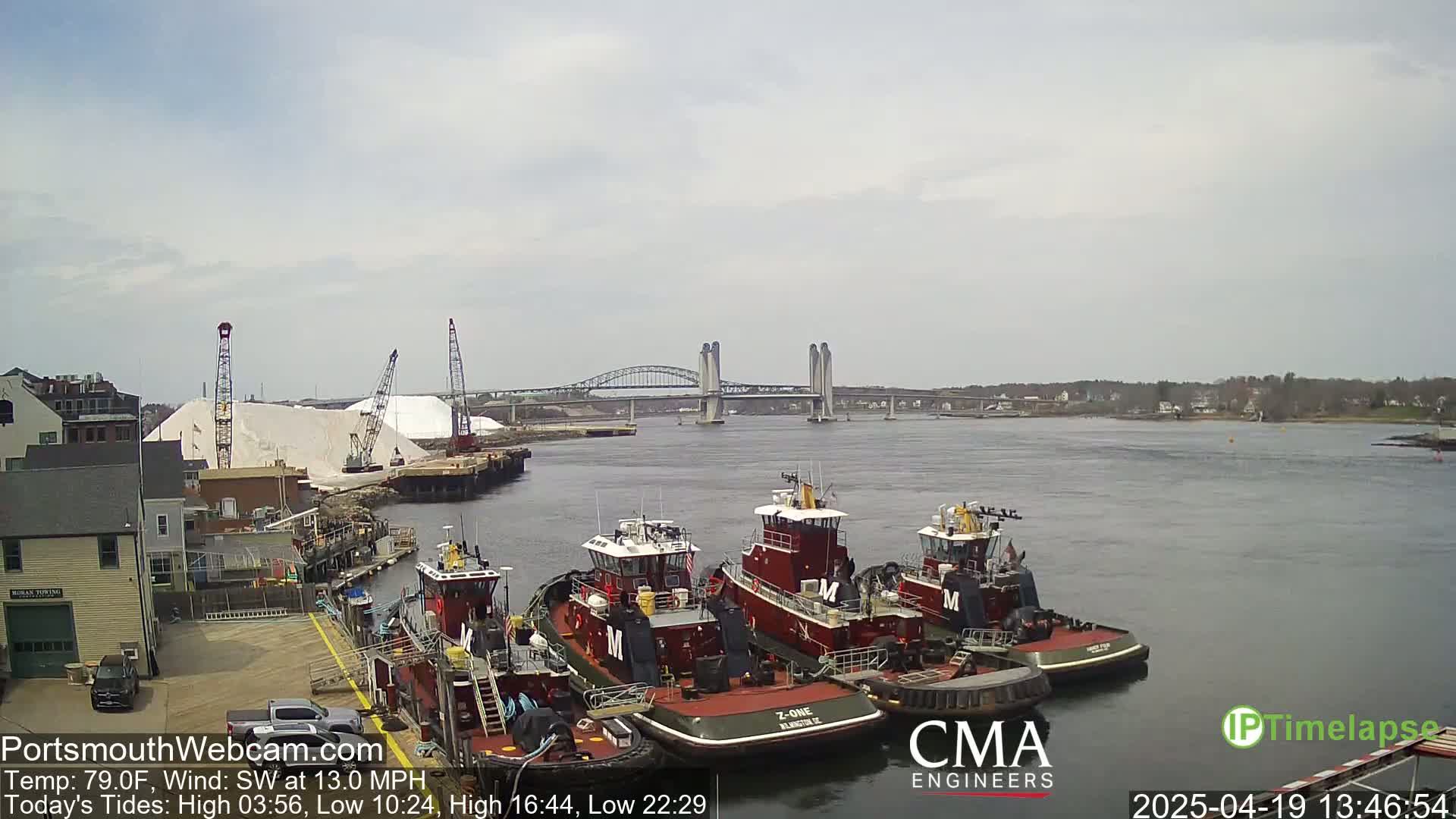 Several tugboats are docked at a waterfront, with a bridge and a large pile of white material visible in the background under a partly cloudy sky.