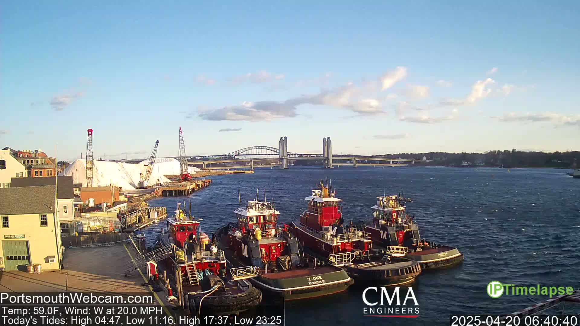 A harbor scene with several tugboats docked, a large pile of salt, and a long bridge in the background under a partly cloudy sky.