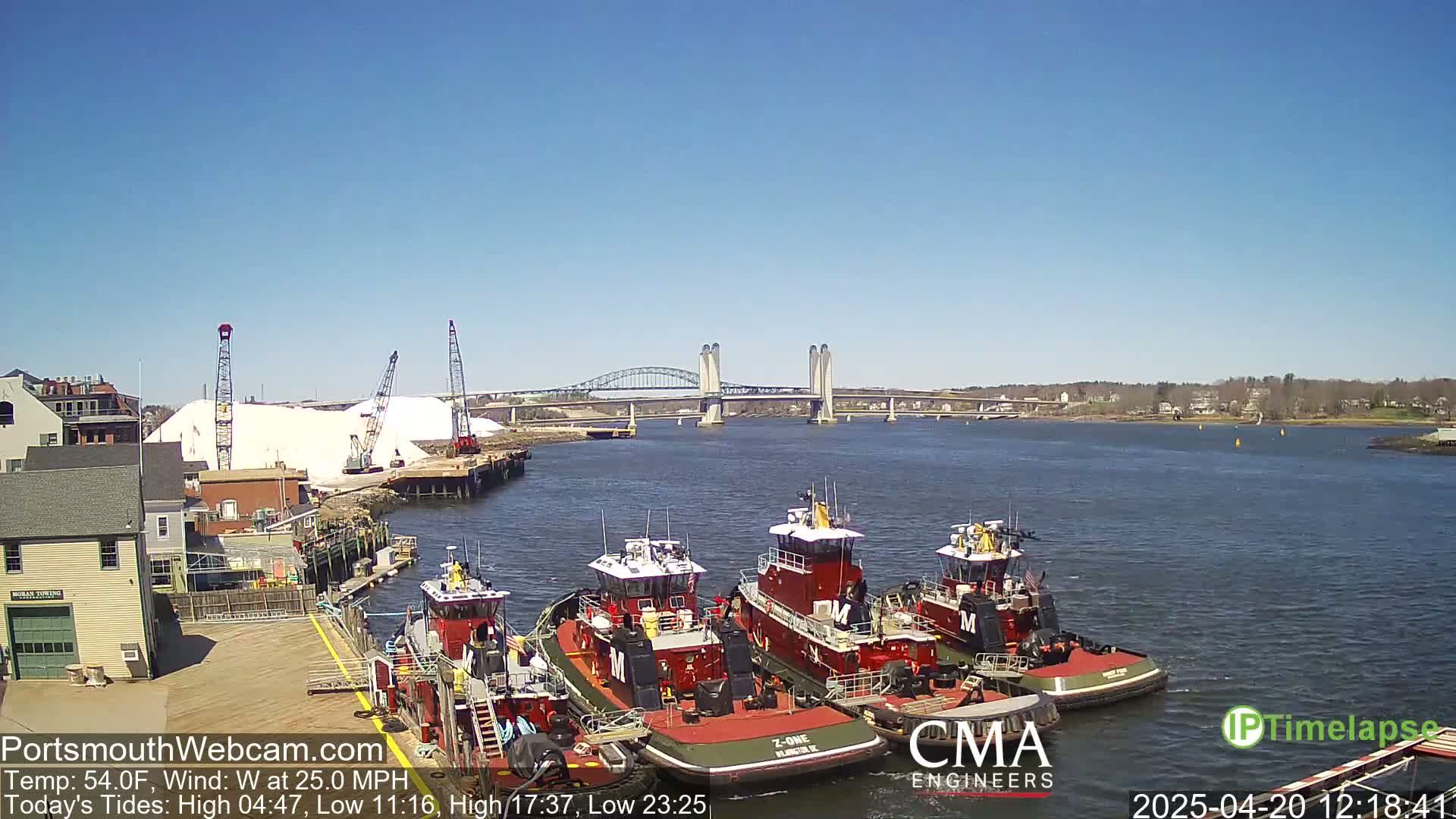 Several tugboats are moored at a dock next to a large pile of white material, with a bridge spanning a waterway under a clear blue sky.