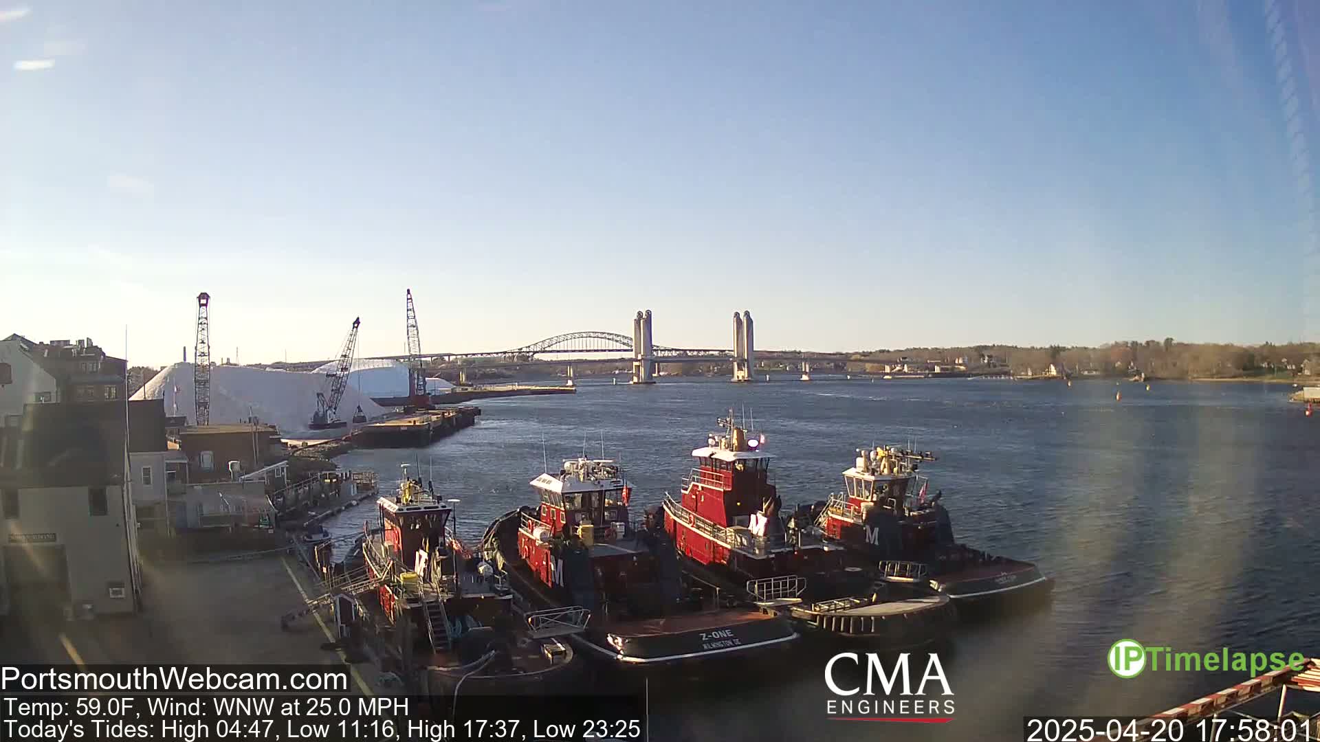 Several tugboats are moored at a dock near a large pile of salt, with a bridge spanning a calm waterway in the background under a clear sky.