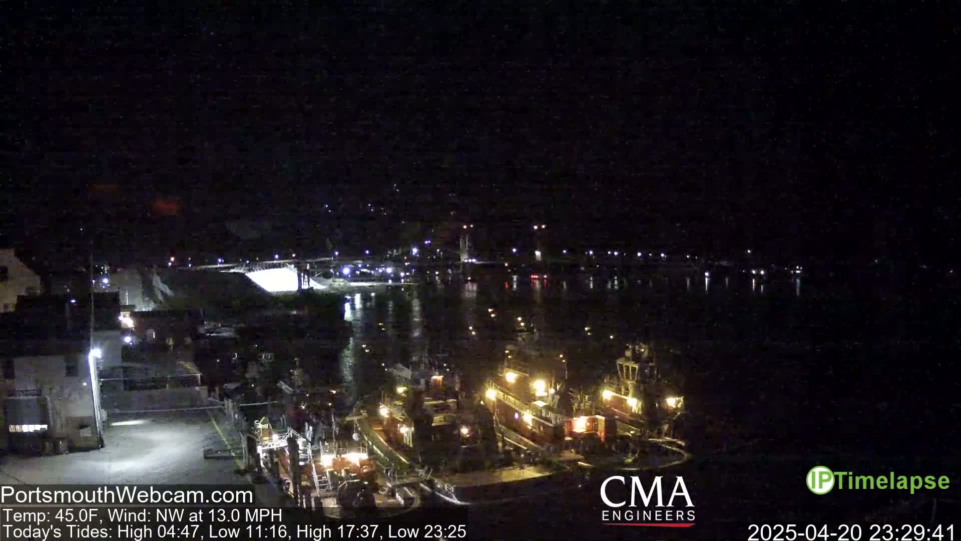 At night, under a dark sky with a northwest wind at 13 mph, several tugboats are docked at a waterfront location near a lit-up roadway.