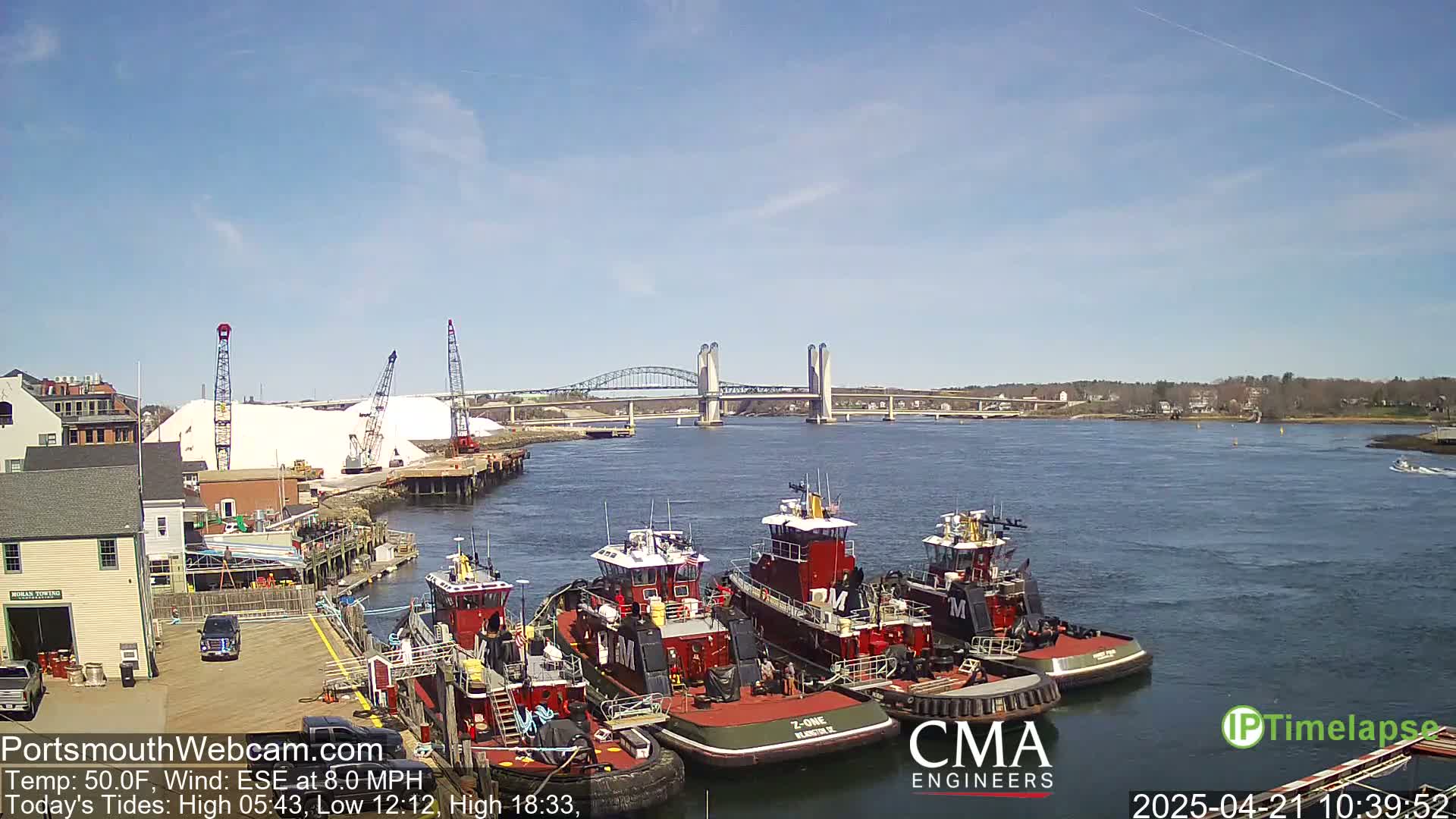 Several tugboats are docked at a waterfront, with a bridge and a large pile of salt visible in the background under a clear, sunny sky.