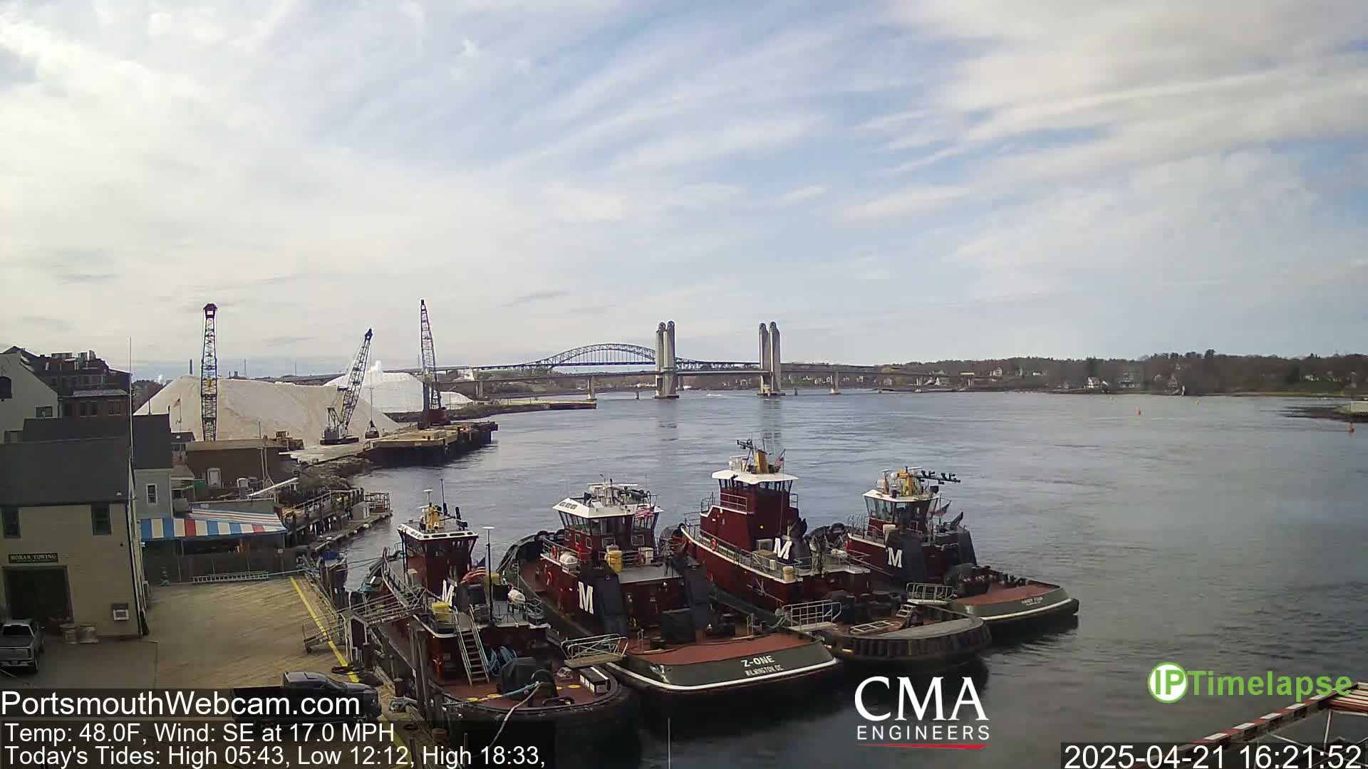 Several tugboats are moored at a dock near a bridge and a pile of white material, under a partly cloudy sky with a southeasterly wind.