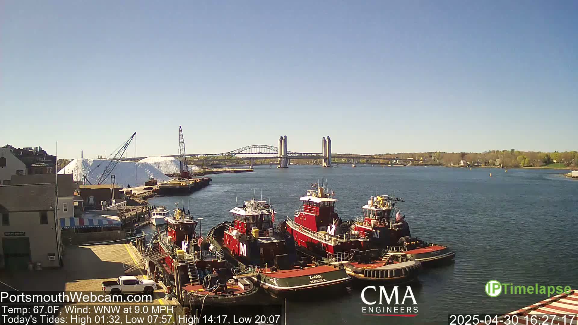 Several tugboats are moored at a dock near a bridge on a sunny day with a light wind.