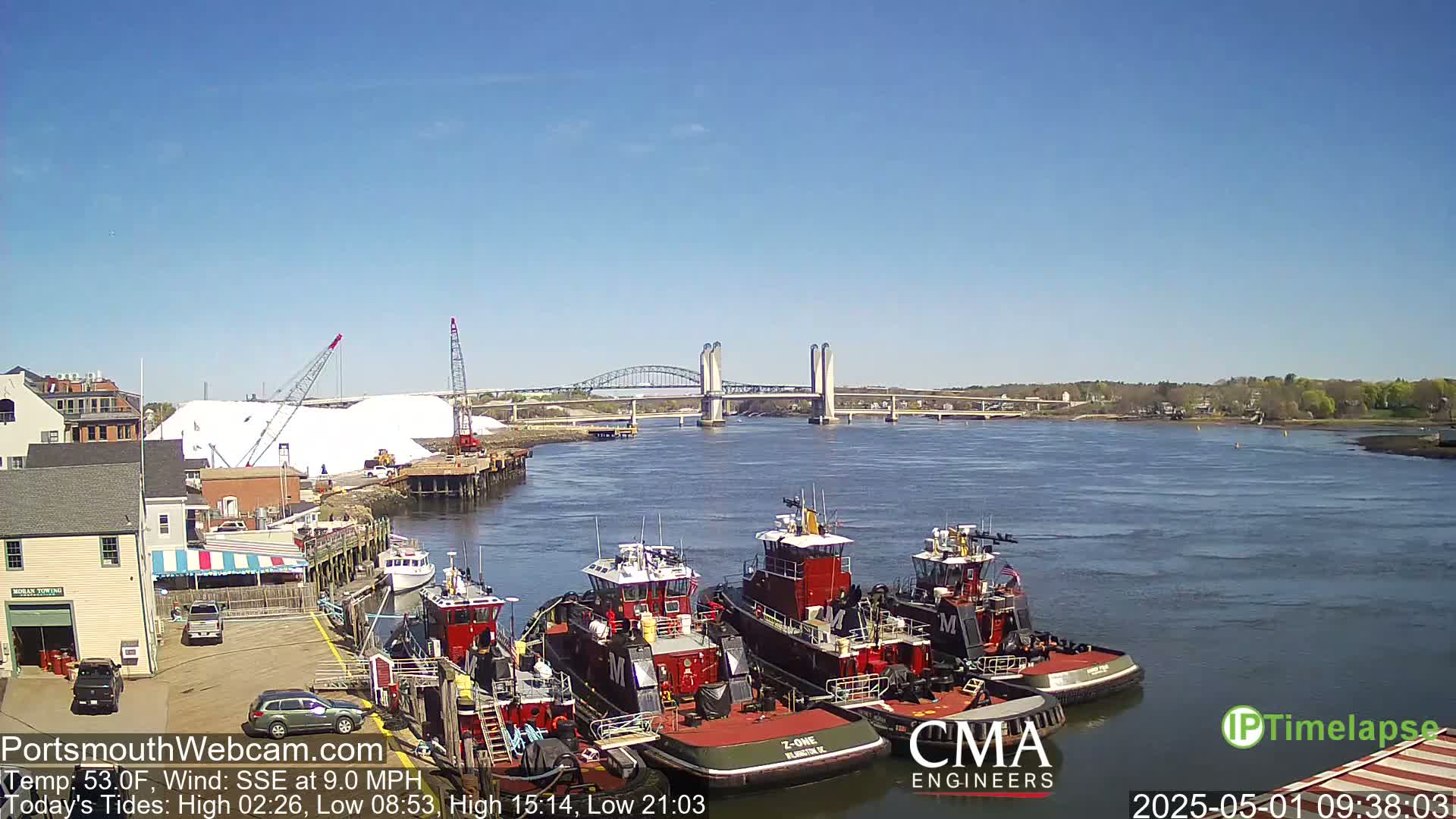 Several tugboats are moored at a waterfront dock near a bridge on a sunny day with a calm river.