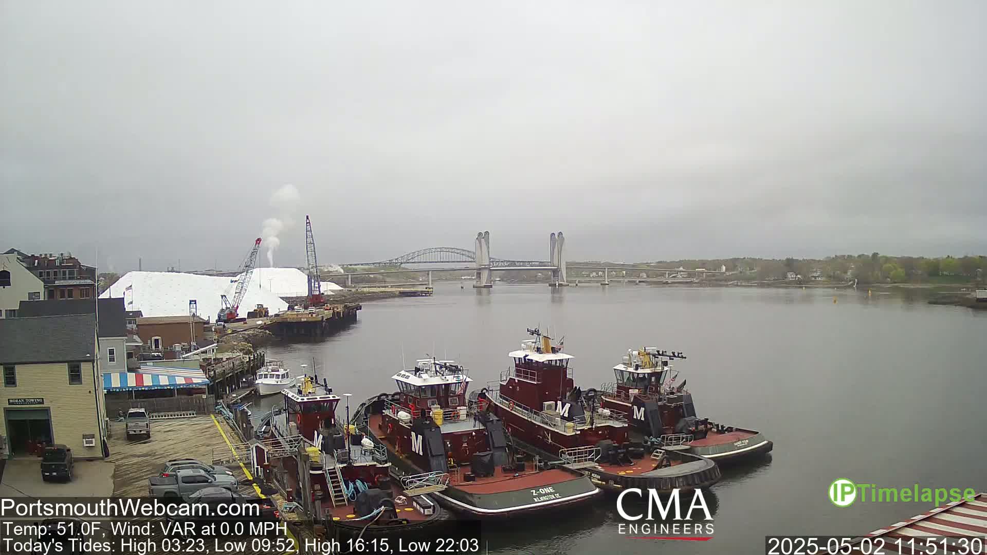 Several tugboats are moored at a dock next to a large pile of white material, with a bridge spanning a calm body of water under an overcast sky.