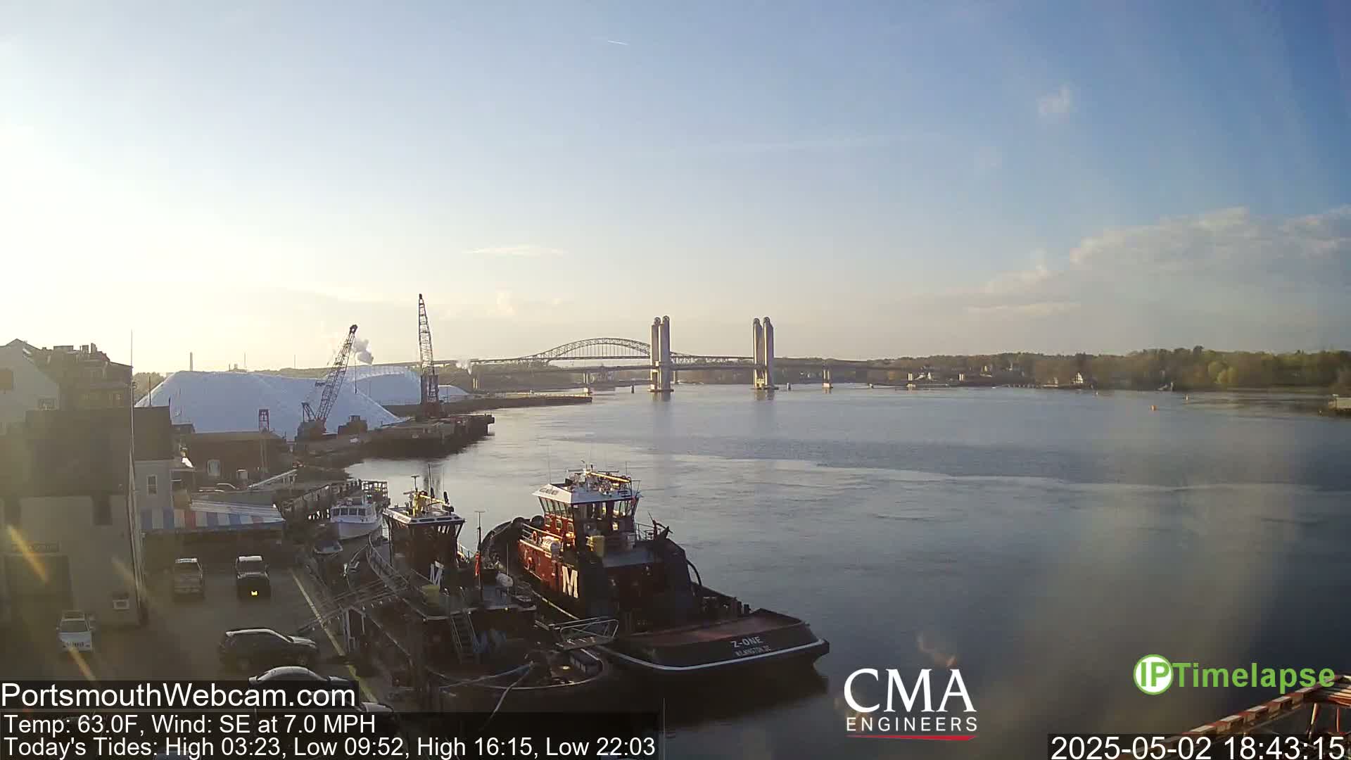 Several tugboats are moored at a waterfront dock, under a partly cloudy sky, with a double-leaf bascule bridge spanning a wide river in the background.