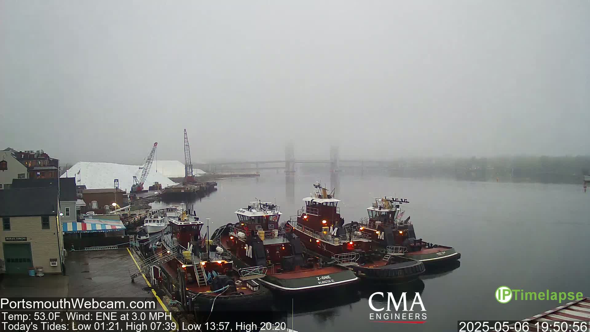 Several tugboats are docked at a waterfront, under foggy conditions, with a large pile of white material and a bridge visible in the distance.