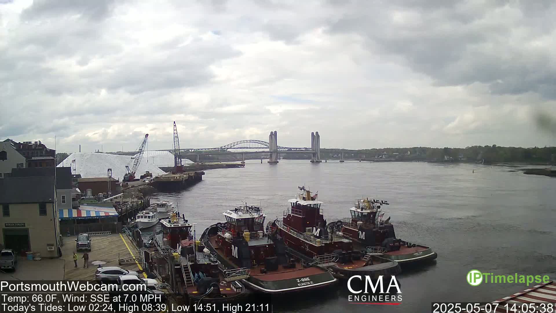 Several tugboats are moored at a dock next to a large pile of white material, with a bridge spanning a river under an overcast sky.