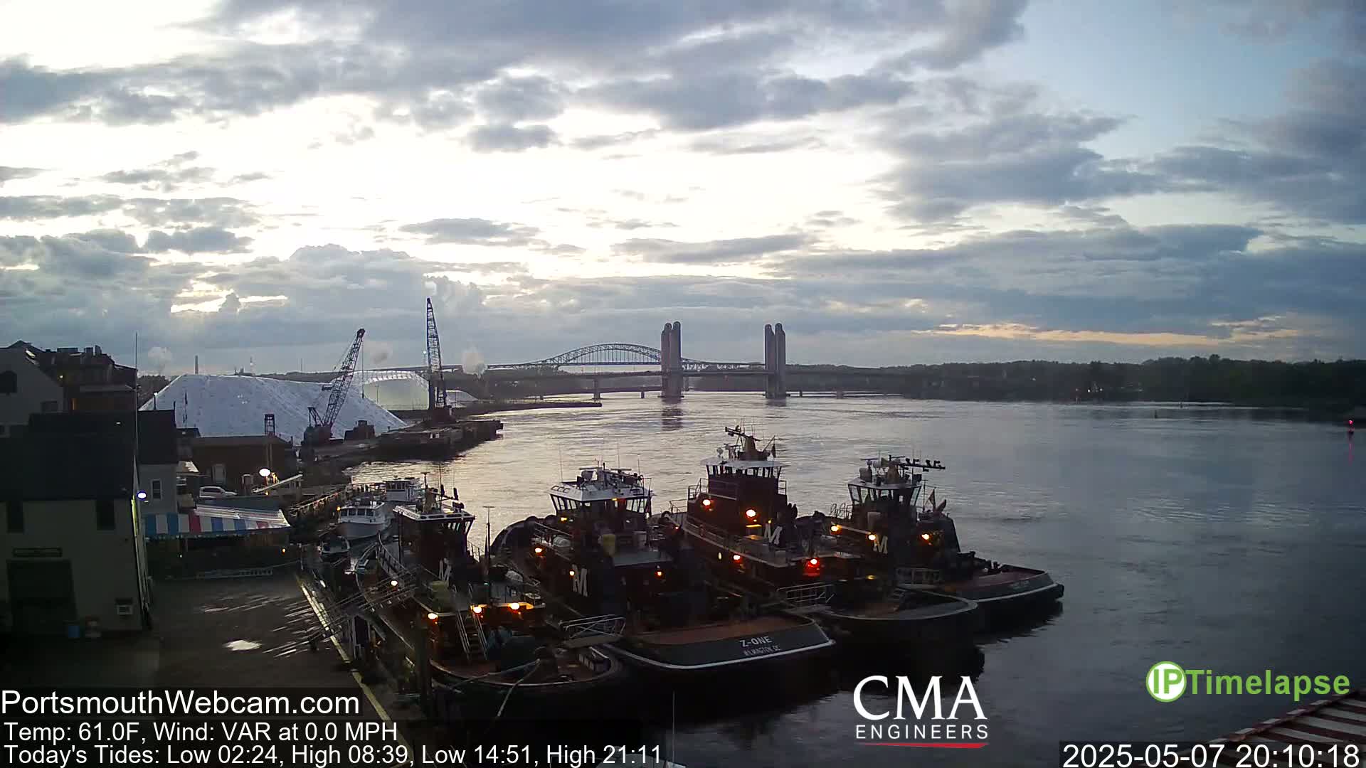 Several tugboats are moored at a dock, under a partly cloudy sky, with a bridge in the background.