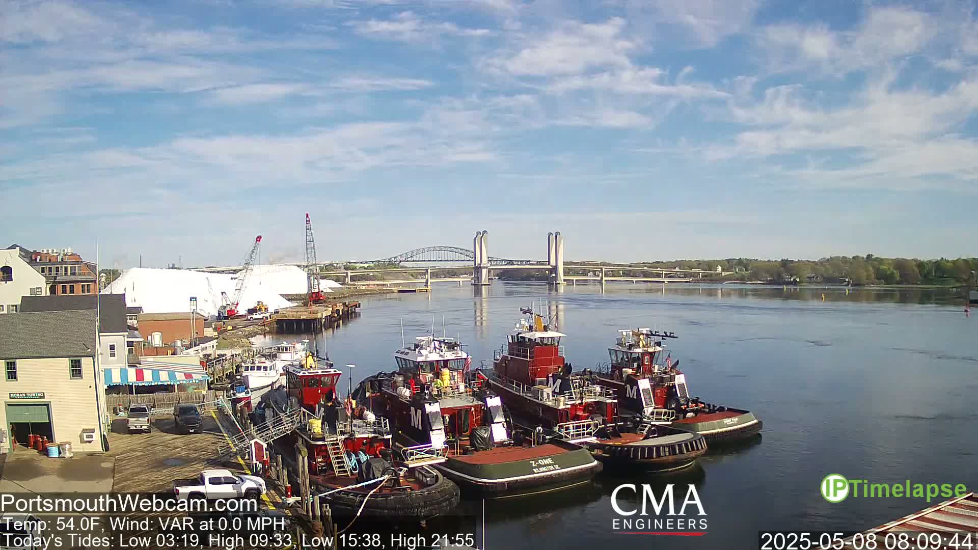 Several tugboats are moored along a waterfront, with a bridge spanning a river under a mostly sunny sky.