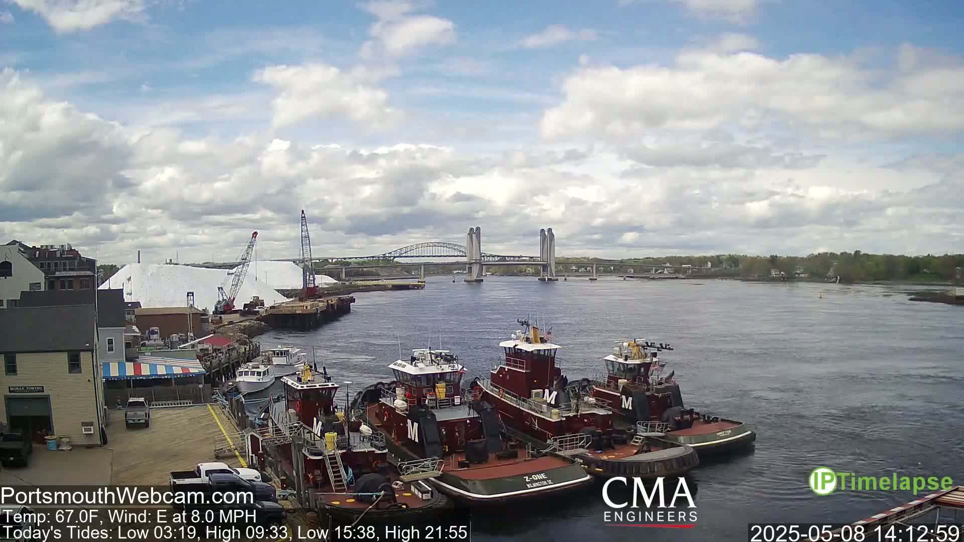Several tugboats are moored at a dock next to a large pile of white material, with a bridge spanning a river in the background under a partly cloudy sky.