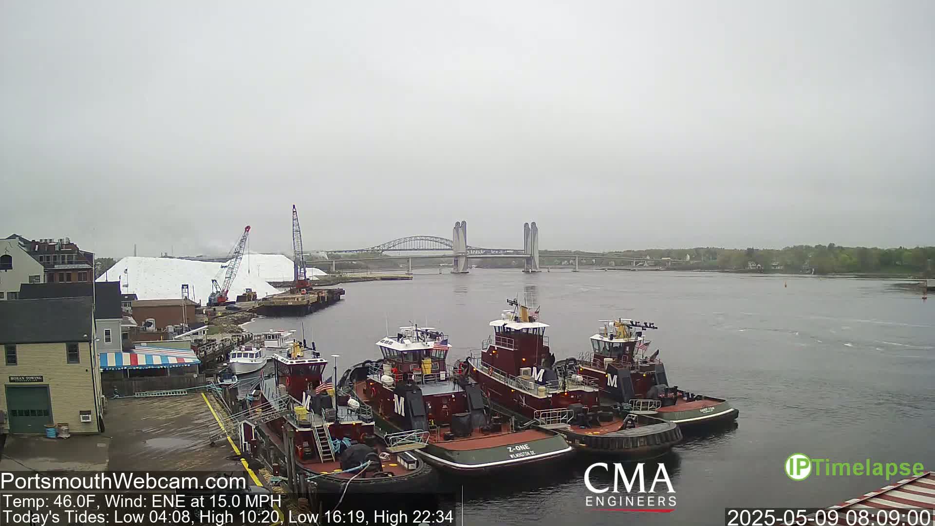 Several tugboats are moored at a dock near a large pile of white material, with a bridge in the distance under an overcast sky.