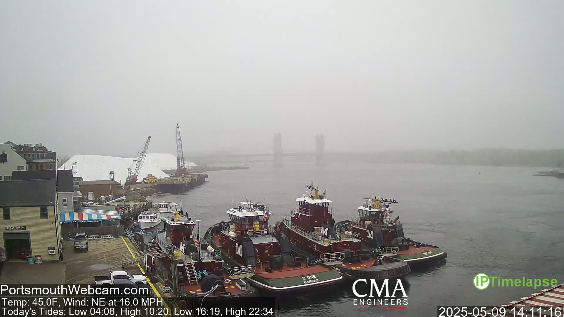 Several tugboats are docked at a wharf in foggy conditions, with a distant bridge barely visible through the haze.