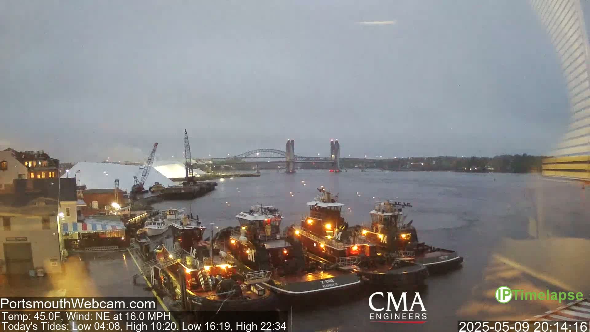 Several tugboats are docked near a waterfront under an overcast sky with a northeast wind of 16 mph.