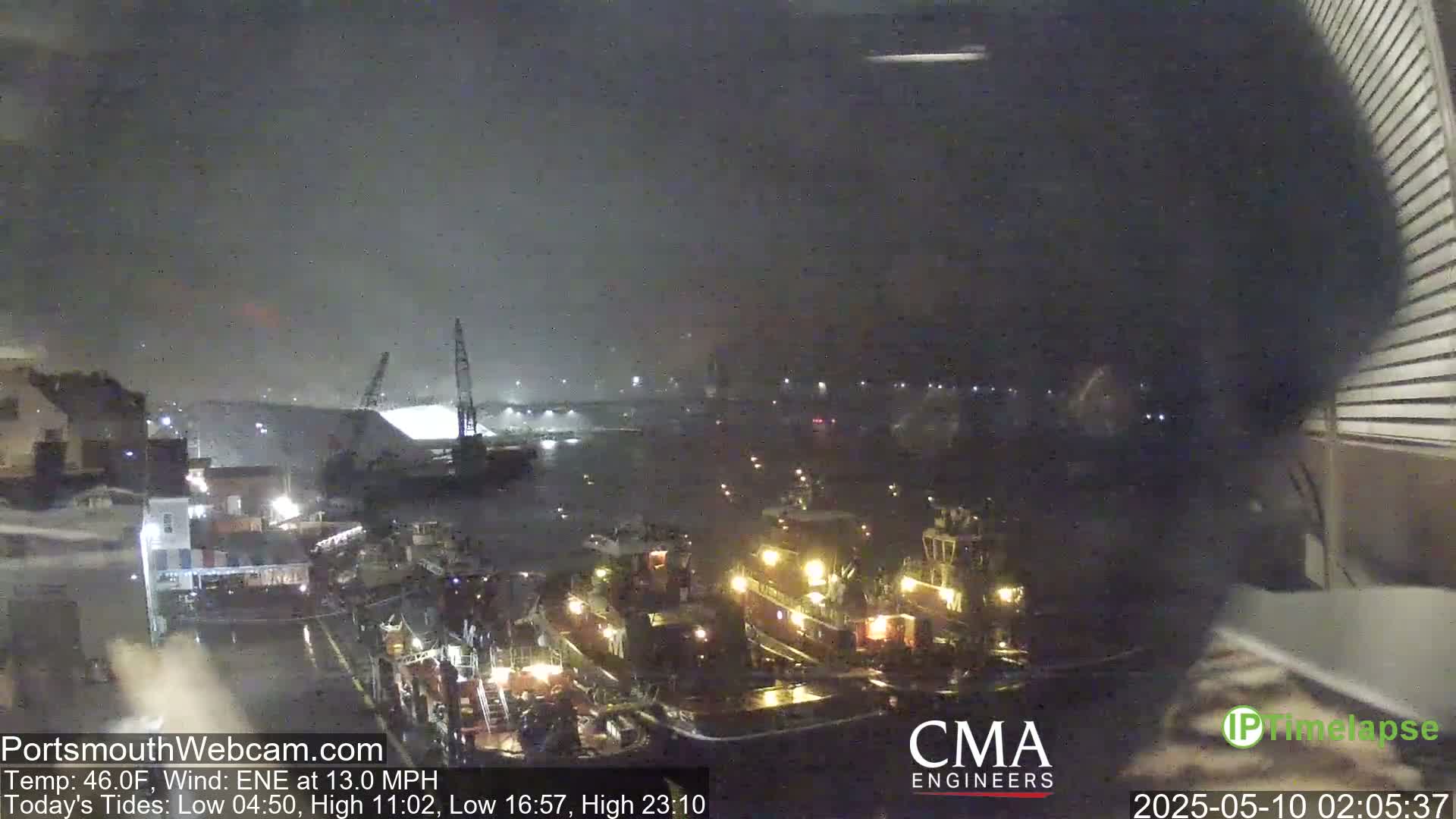 At night, under a dark, somewhat hazy sky, several tugboats are docked in a harbor near a large structure, with cranes visible in the background.
