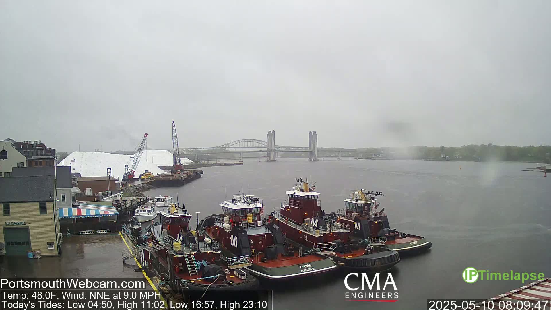 Several tugboats are docked at a waterfront, with a large pile of white material and a bridge visible in the distance under an overcast sky.