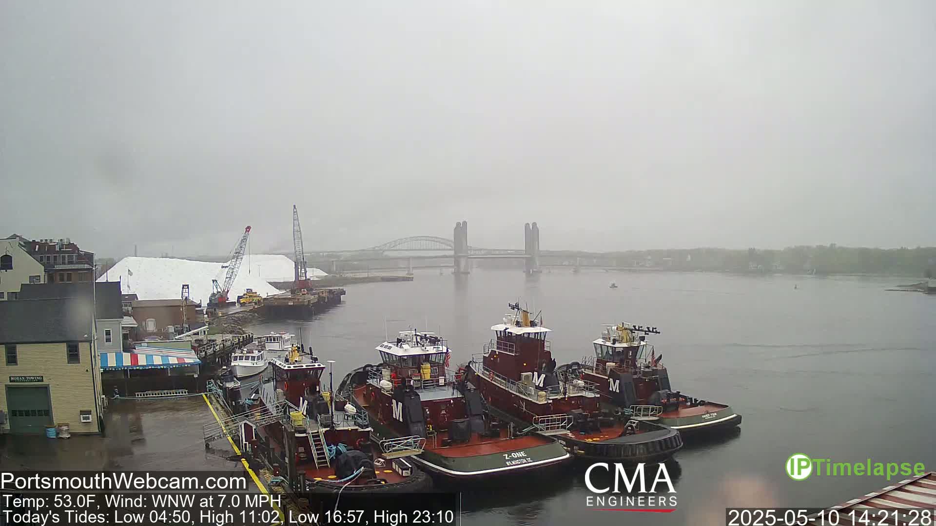 Several tugboats are moored at a dock in foggy conditions, with a bridge visible in the distance.