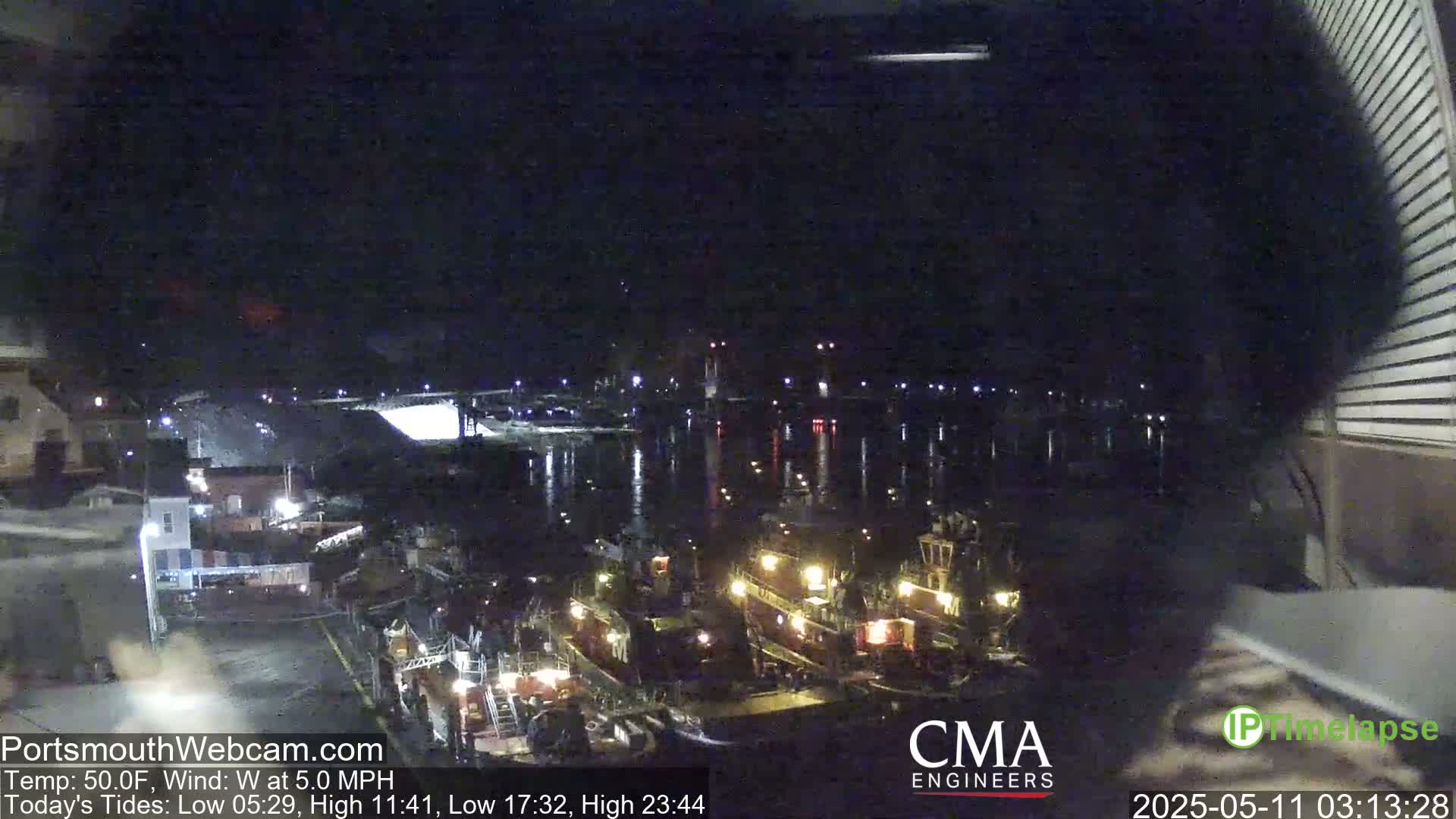 A nighttime view of several tugboats docked at a harbor, with a calm body of water reflecting lights from the shore under a dark sky.
