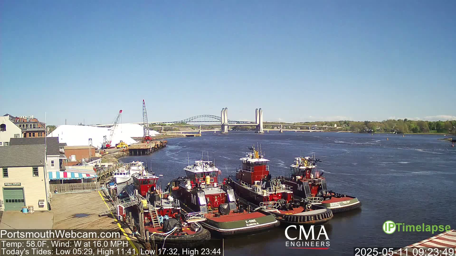 Several tugboats are moored at a dock next to a large pile of white material, with a bridge spanning a river in the background under a clear blue sky.