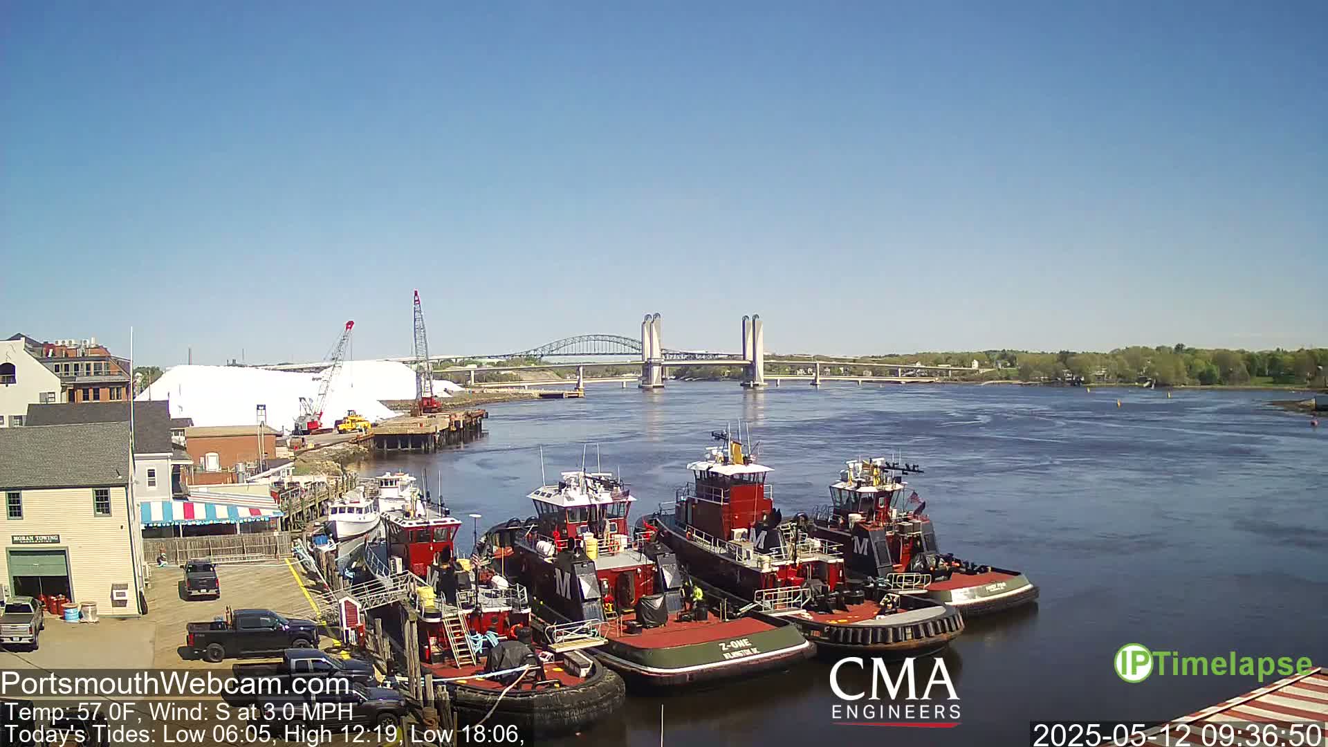 A sunny day reveals several tugboats docked along a waterfront, with a bridge spanning a river in the background and a large pile of white material nearby.