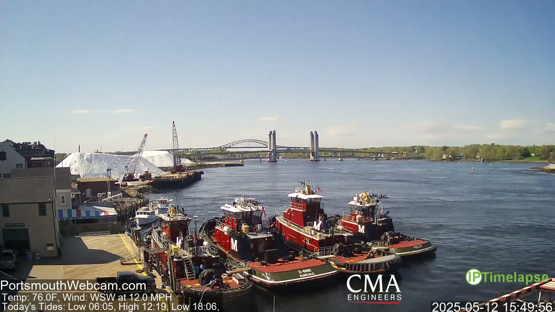 Several tugboats are moored at a dock next to a large pile of white material, with a lift-bridge spanning a calm river in the background under a clear sky.