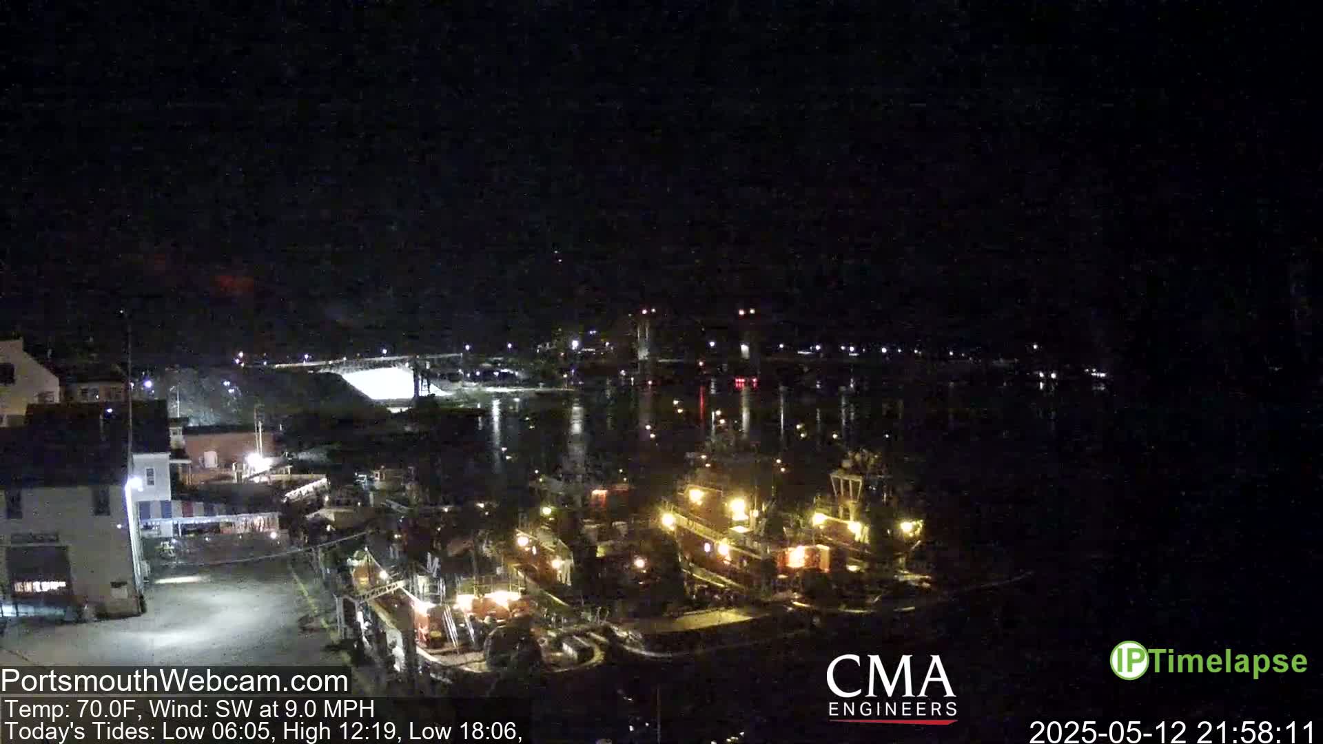 A nighttime harbor scene shows several docked tugboats illuminated, with calm water reflecting the lights of a nearby town under a clear, dark sky.