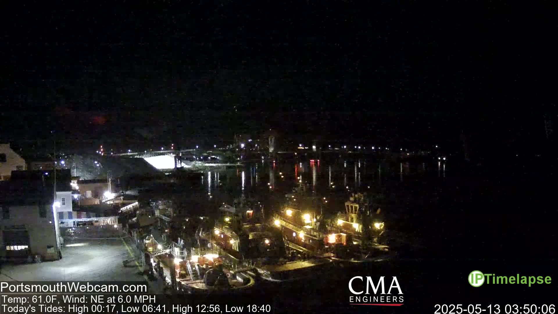 A nighttime harbor scene shows several docked tugboats illuminated, with calm water reflecting the lights under a dark sky.