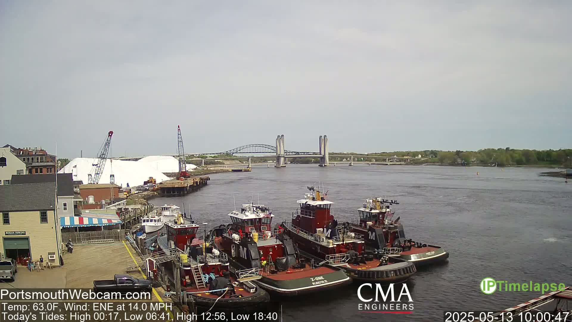 Several tugboats are moored at a dock next to a large pile of white material, with a bridge spanning a calm river in the background under a mostly cloudy sky.