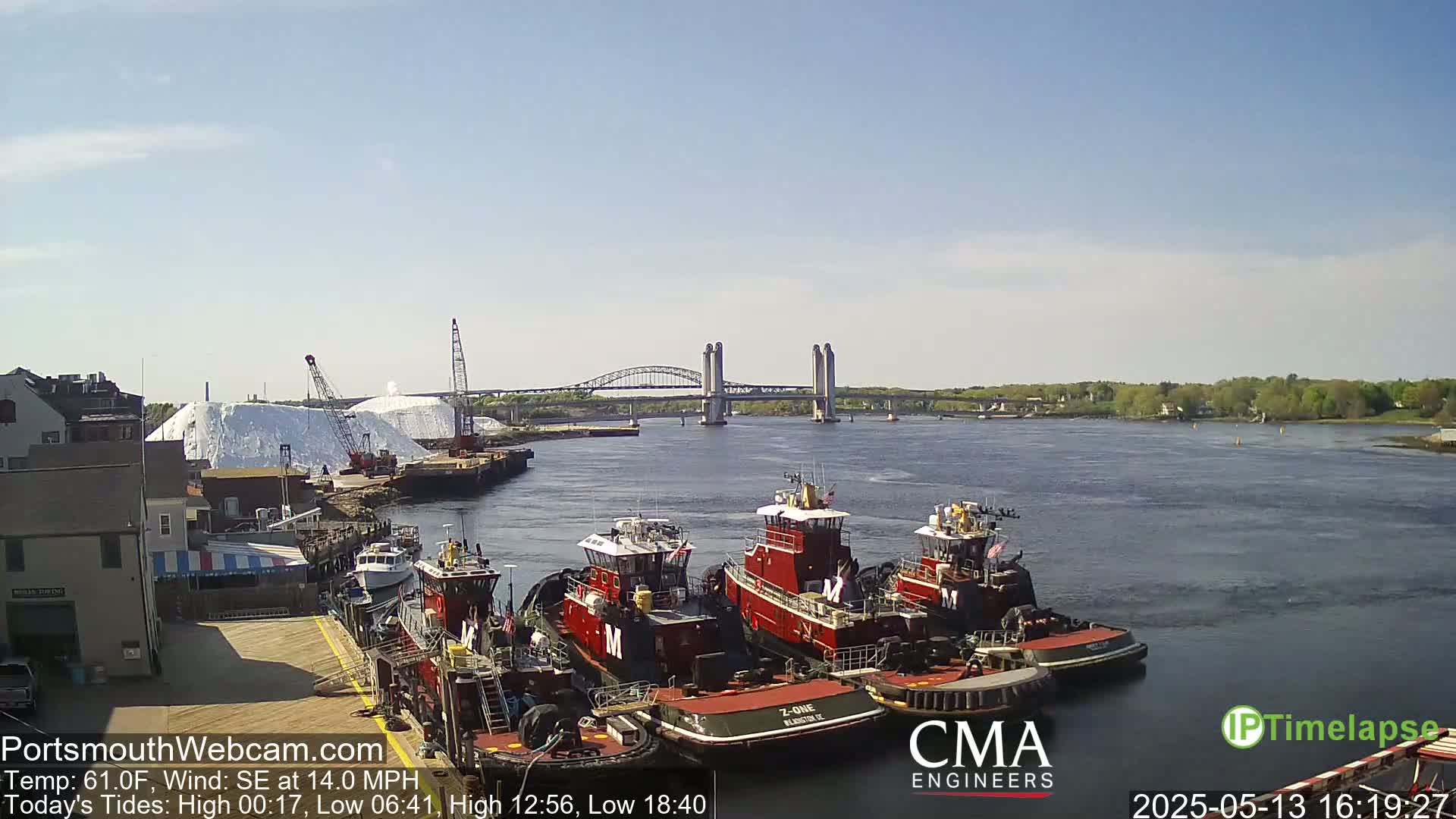 Several tugboats are moored at a dock next to a large pile of white material, with a lift bridge in the background under a clear, sunny sky and a southeast wind.