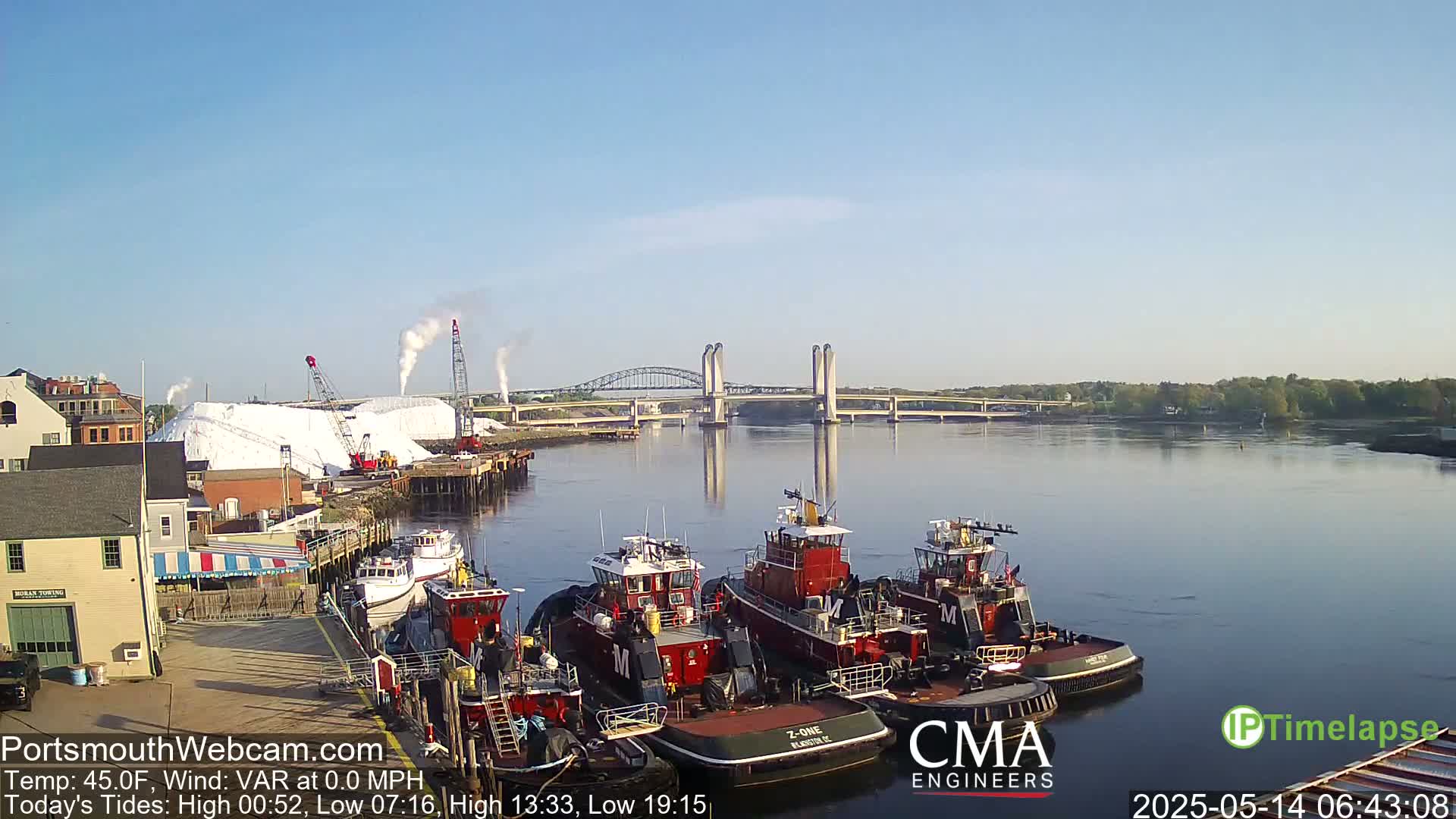 Several tugboats are docked at a waterfront, with a large pile of white material and a lift bridge in the background on a clear, sunny day.
