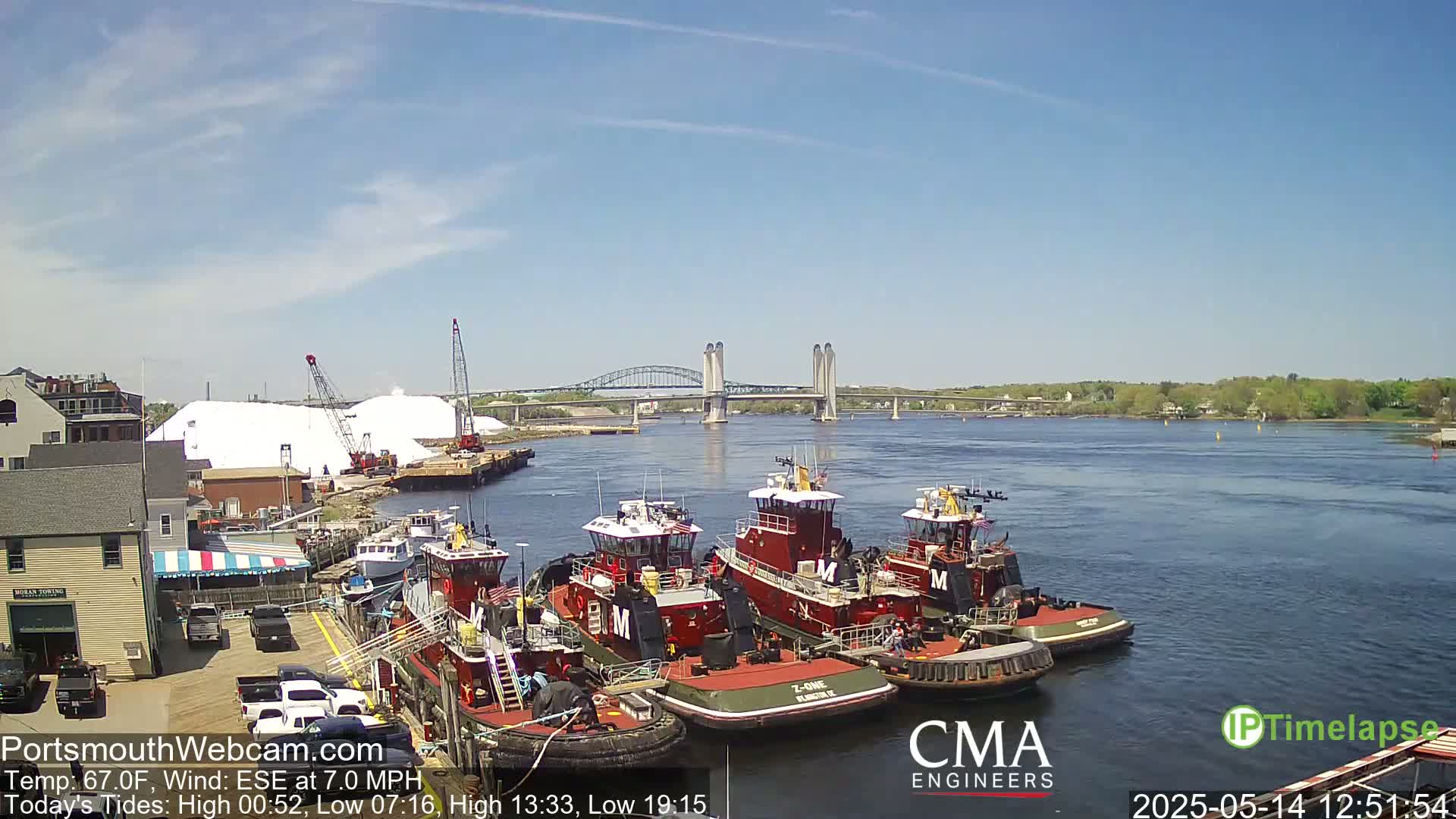 Several tugboats are moored at a waterfront dock, under a sunny sky, with a bridge and a large pile of salt visible in the background.