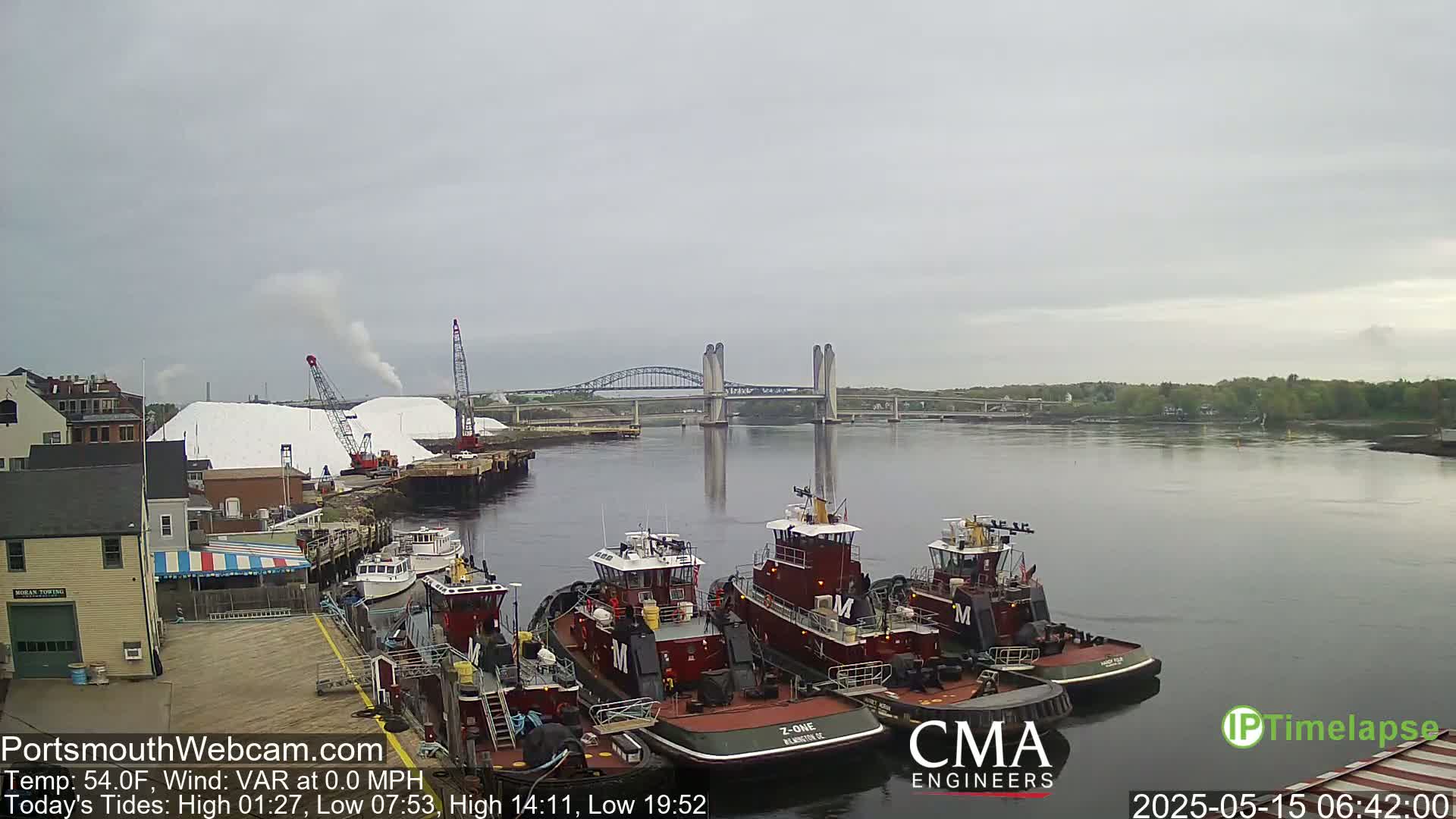 Several tugboats are moored at a dock next to a salt pile and cranes, with a bridge spanning a calm river in the background under an overcast sky.