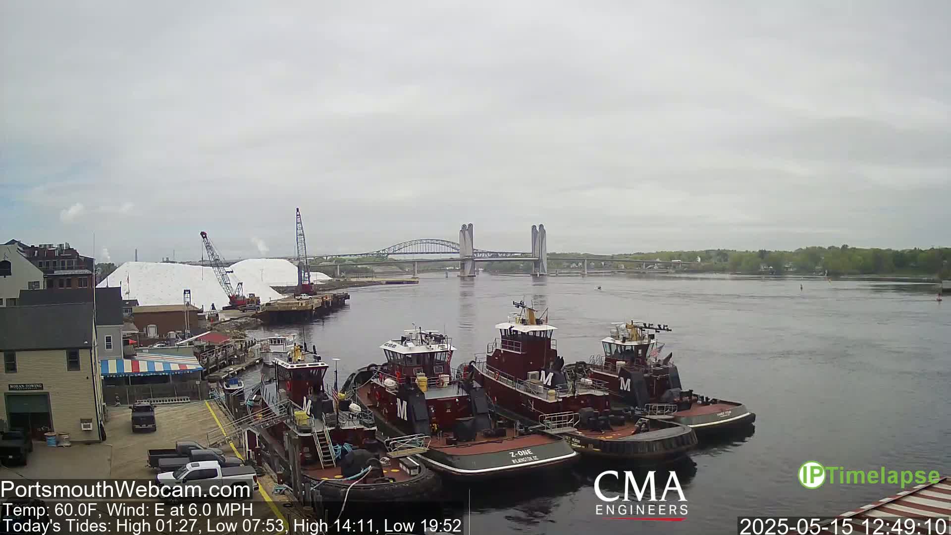 Several tugboats are moored at a dock next to a body of water under an overcast sky, with a bridge in the distance.
