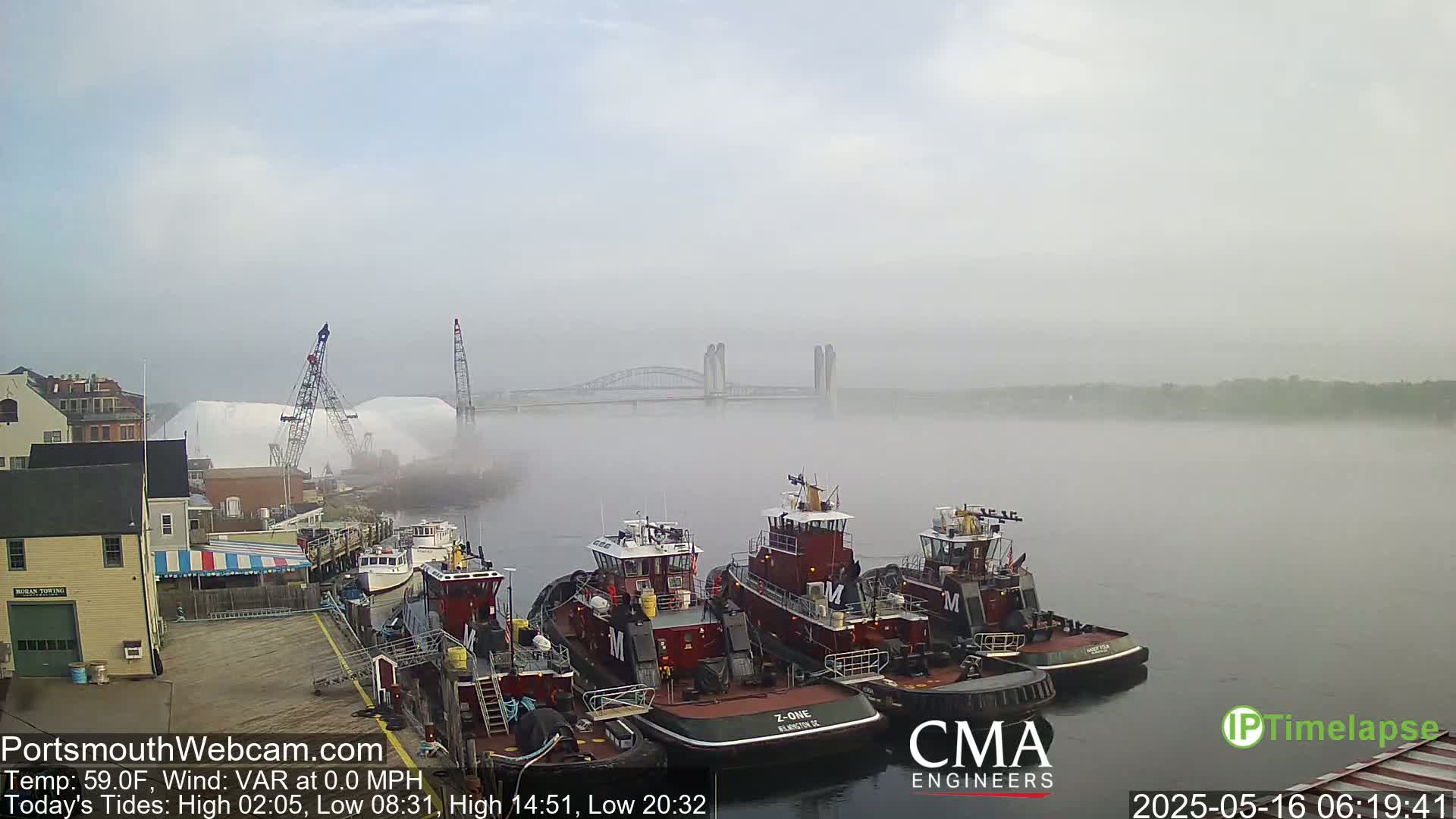 Several tugboats are moored at a dock in a foggy harbor, with a bridge visible in the distance.