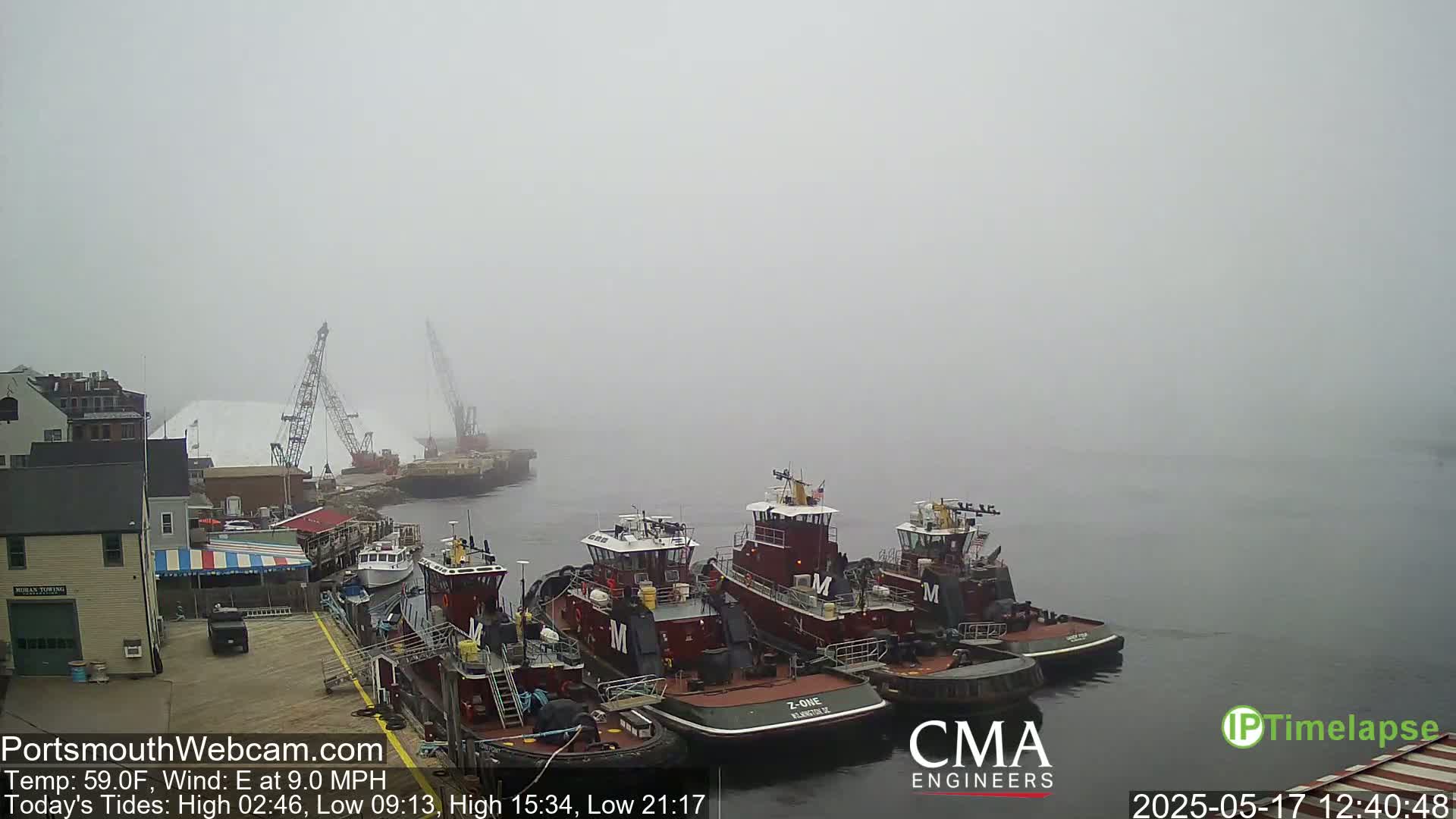 Several tugboats are docked at a wharf in foggy conditions, with a large pile of white material and cranes visible in the background.