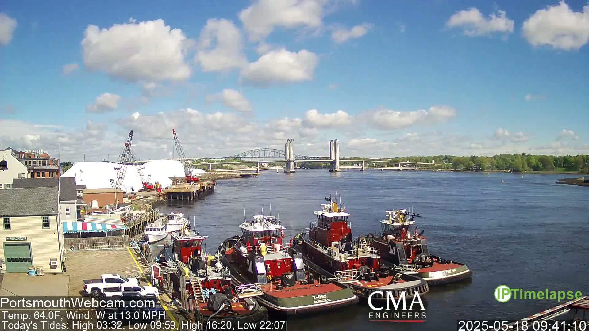 Several tugboats are moored at a dock next to a large pile of salt, with a bridge spanning a river in the background under a partly cloudy sky.