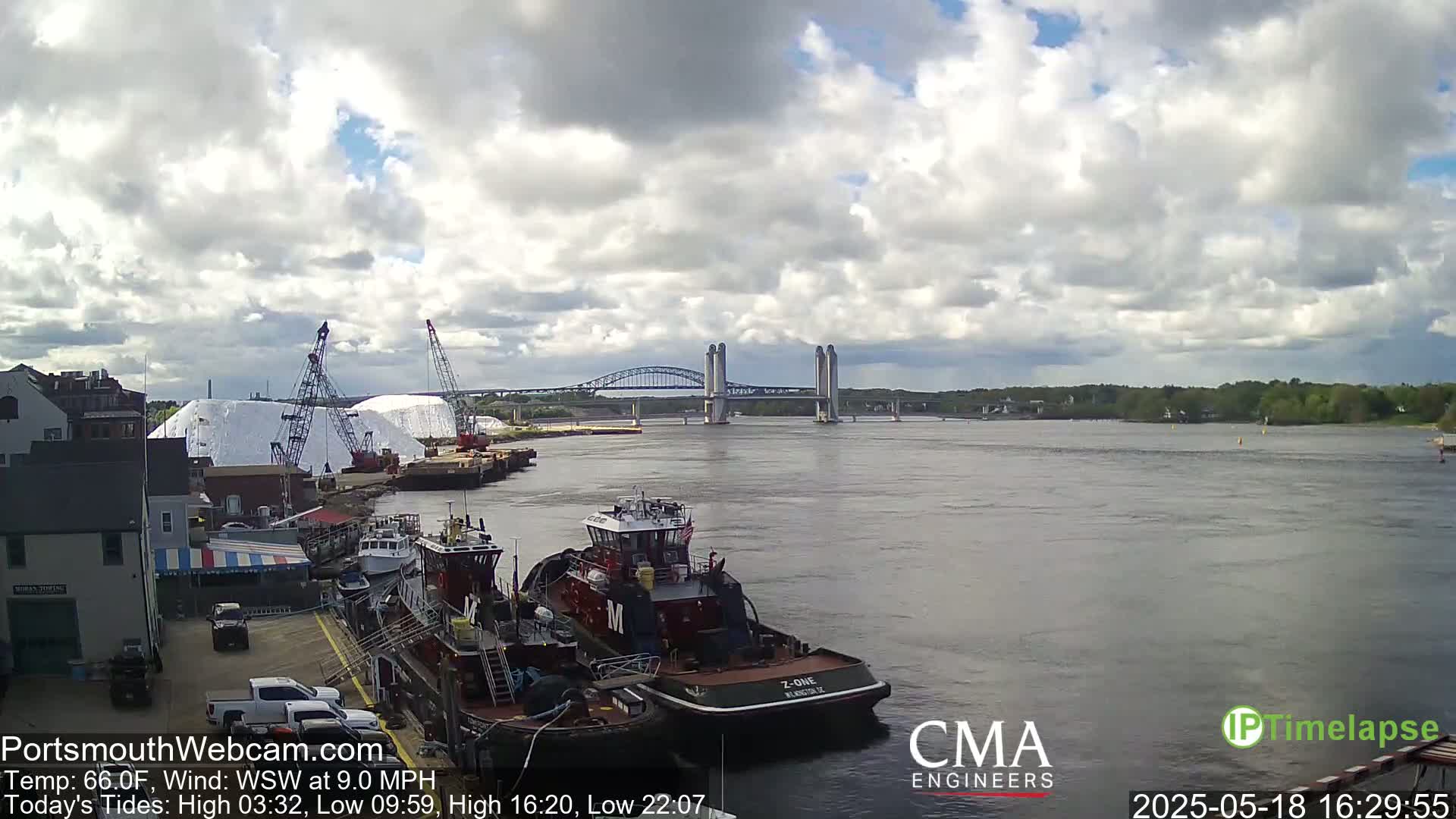 A partly cloudy day shows a river with several tugboats docked near a salt pile and cranes, with a double-decker bridge in the distance.