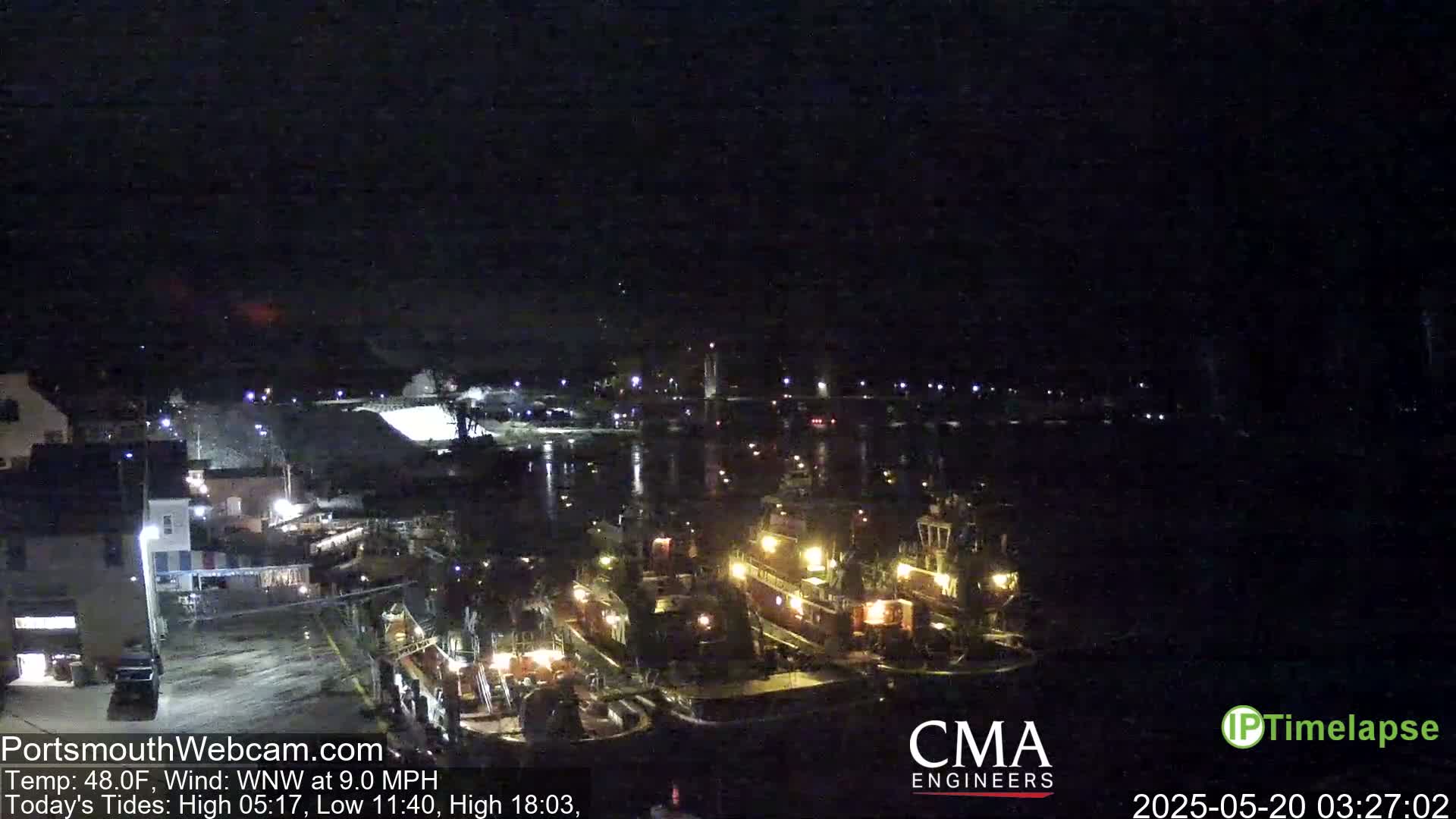 A nighttime view of a harbor with several illuminated tugboats and other vessels docked, under a clear, dark sky.